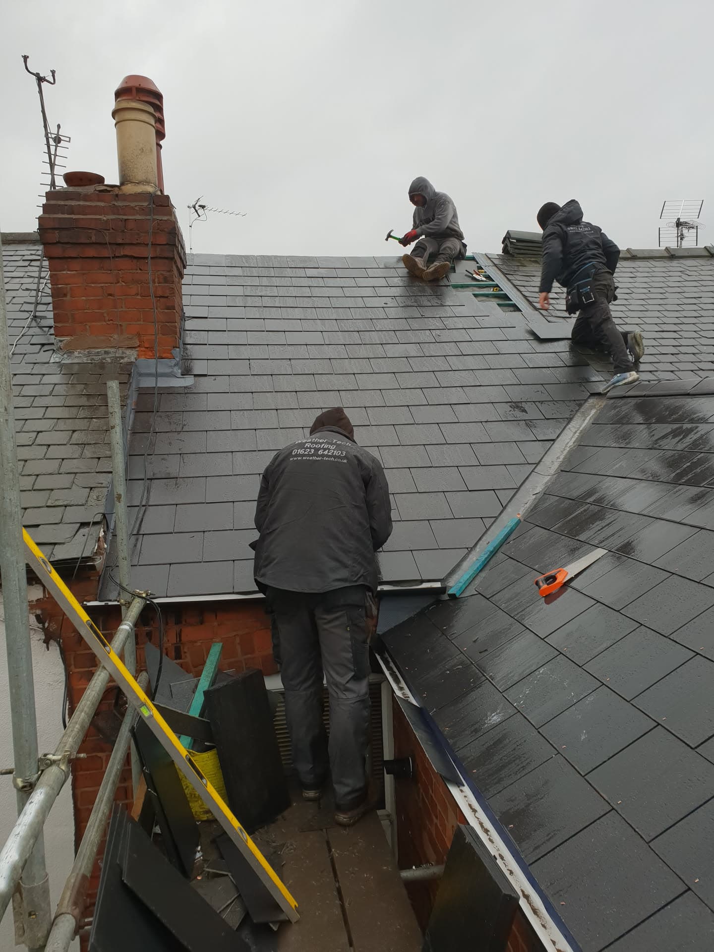 Three fitters installing grey slate tiles on a pitched roof with brick chimney stack