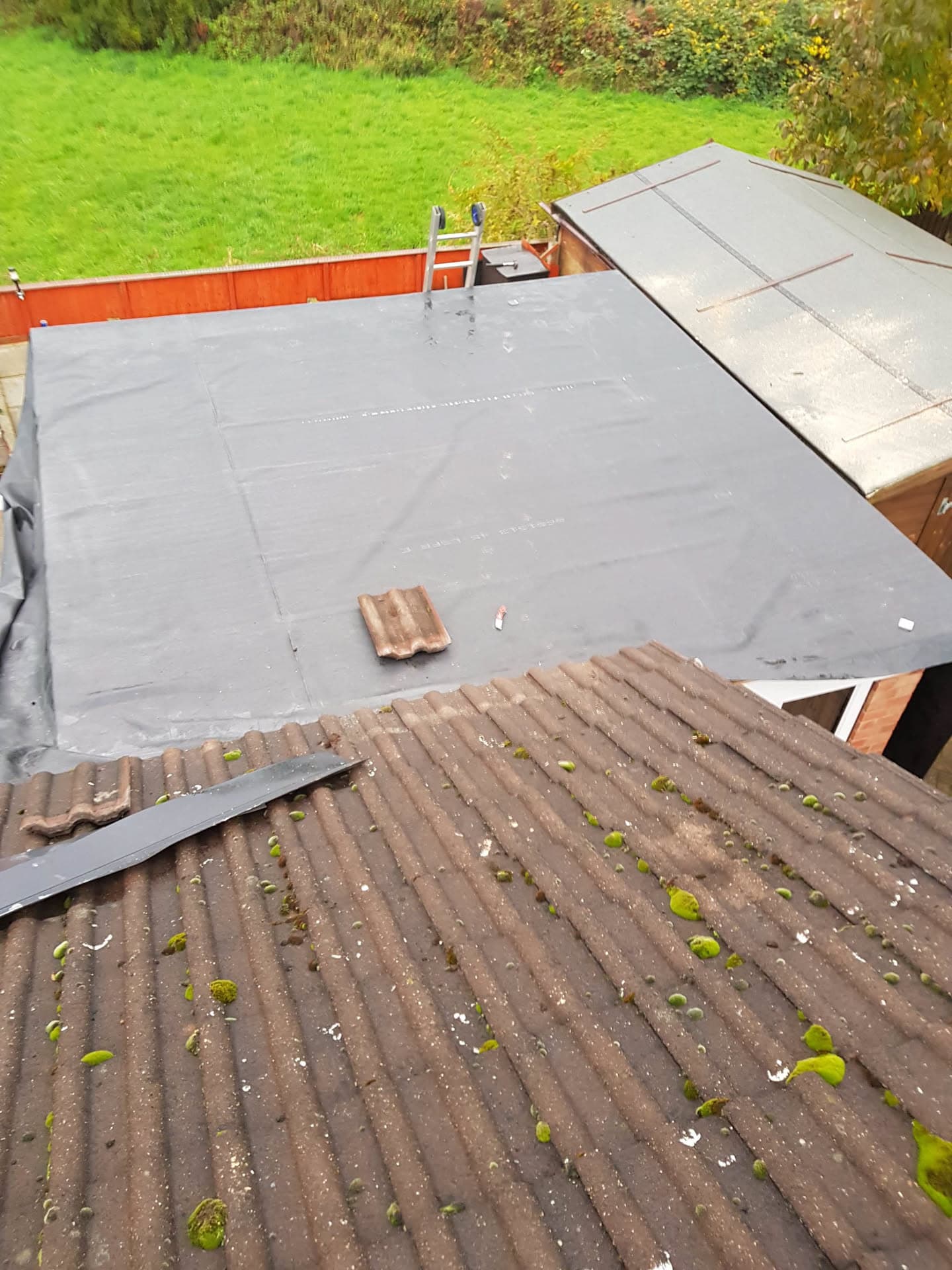 Grey flat roof installation in progress with ladder and adjacent corrugated roof section