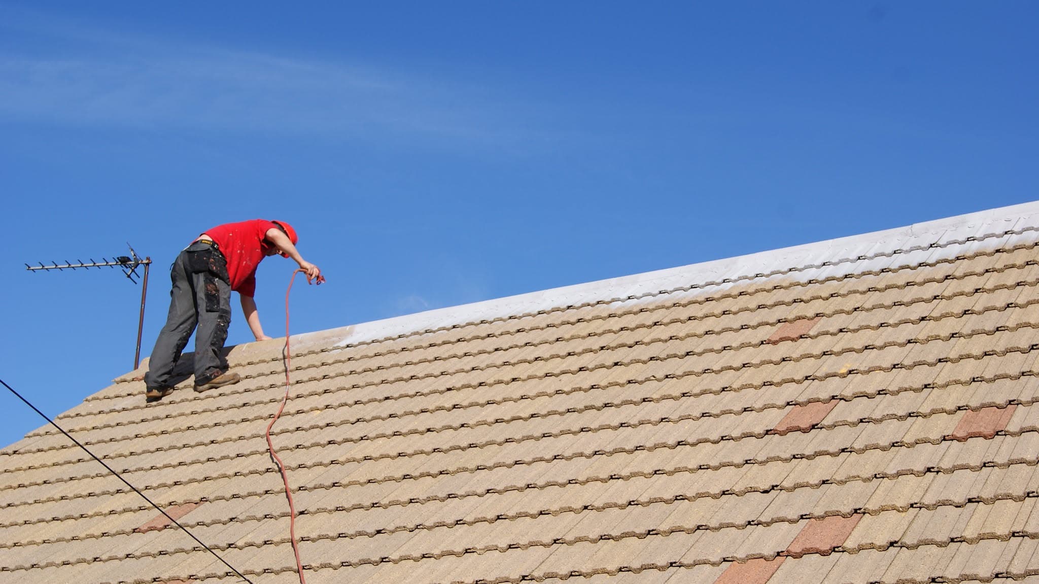Roofer in red shirt working on pitched concrete tiled roof with ridge visible