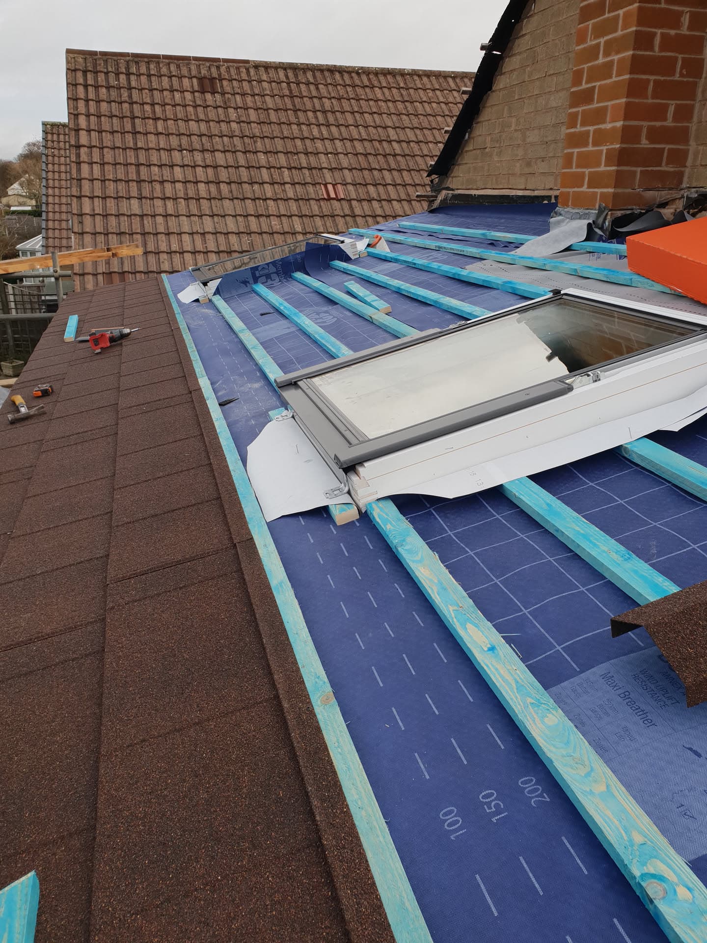 Flat roof installation in progress with blue membrane and velux window on brown tiled pitched roof