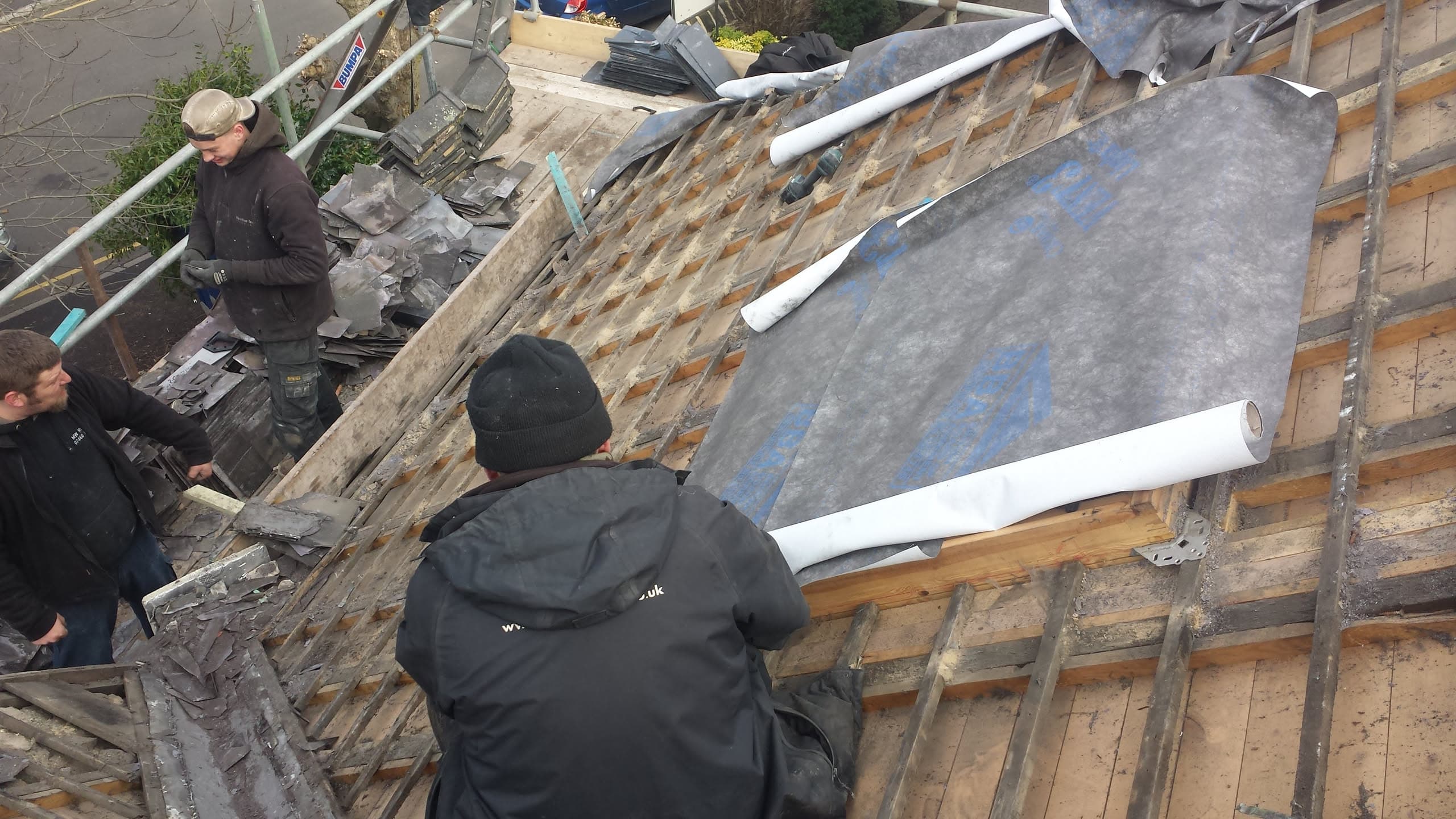 Fitters installing grey felt membrane on pitched roof with exposed timber battens and scaffolding
