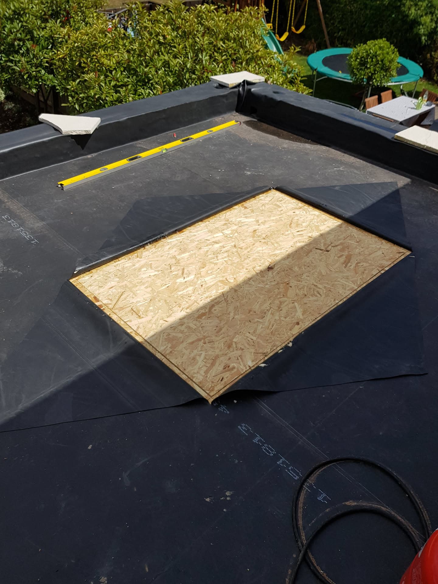 Flat roof installation in progress with black felt membrane and exposed OSB board around skylight opening