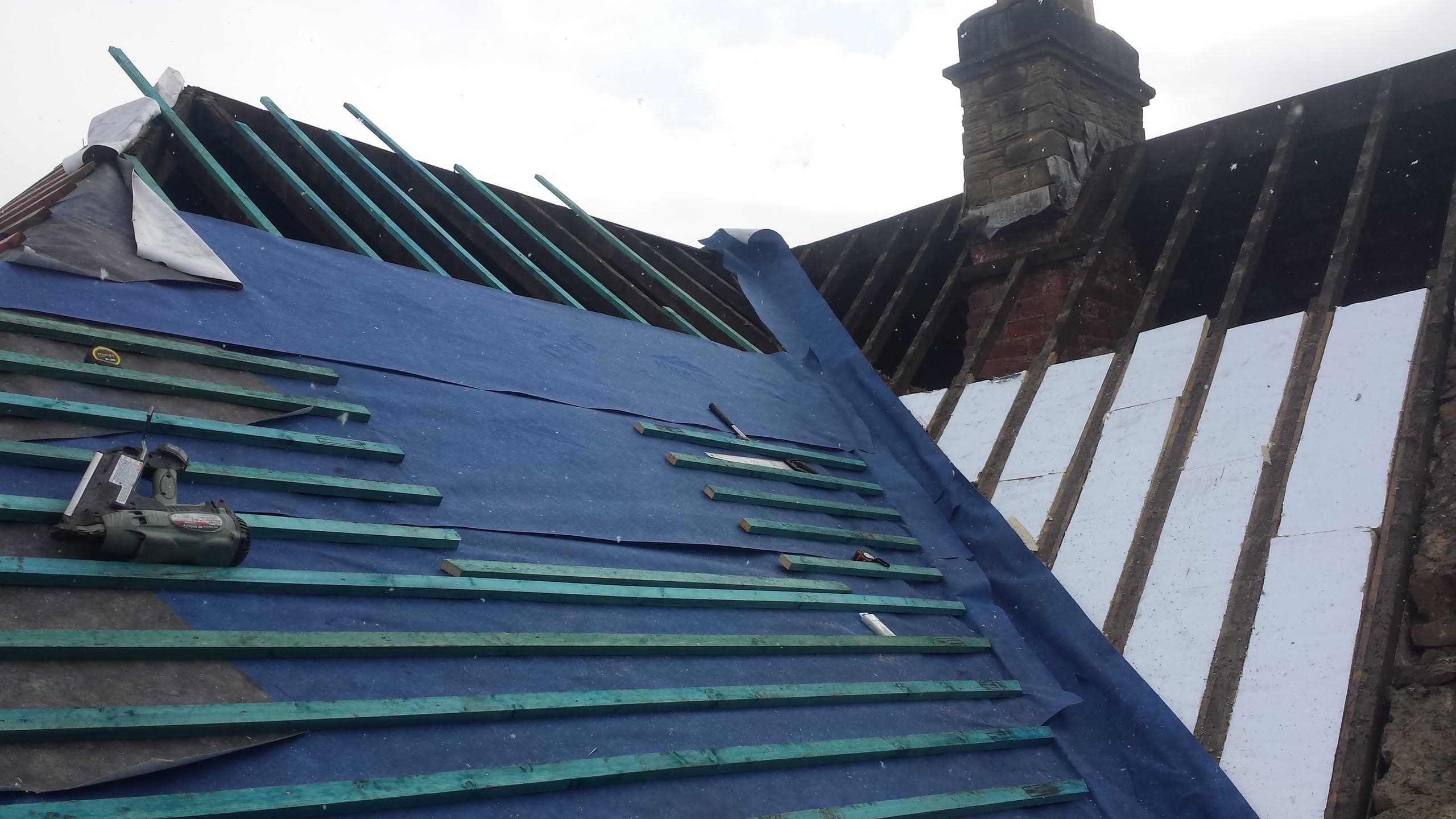Pitched roof with dark green metal sheeting being installed, brick chimney visible
