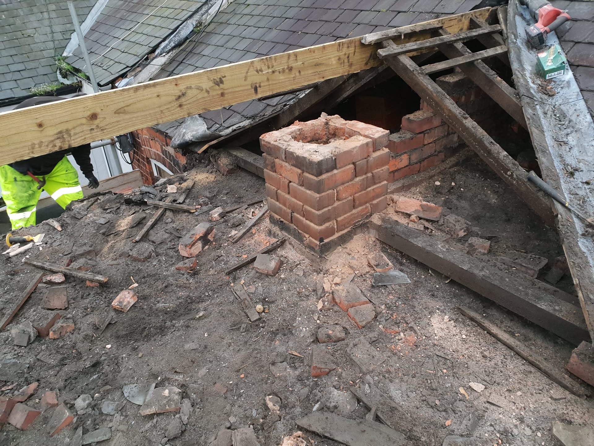 Chimney stack rebuild in progress on pitched slate roof with exposed timber framework and fitter visible