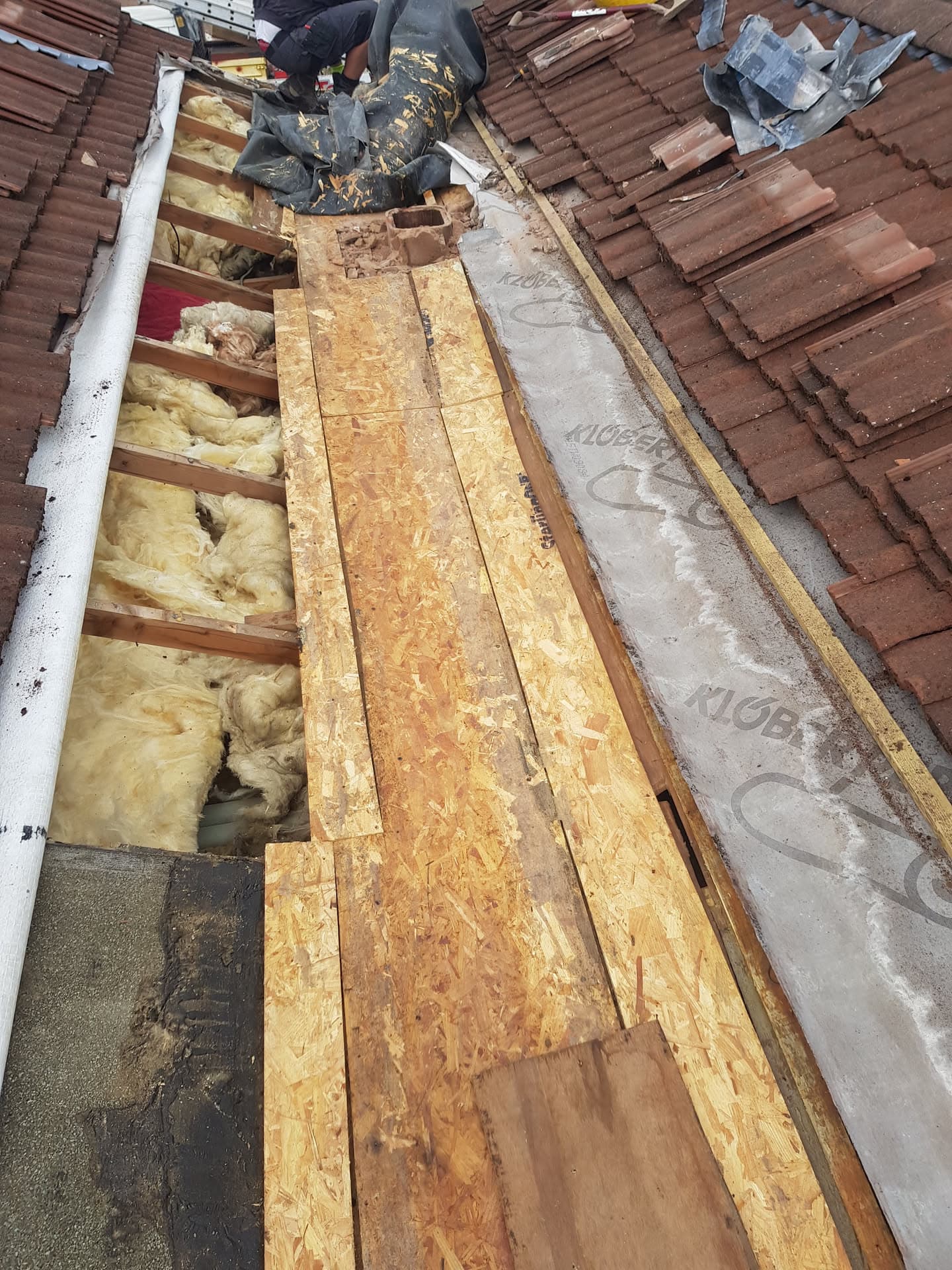 Fitter repairing pitched roof with exposed timber rafters, insulation and brown concrete tiles