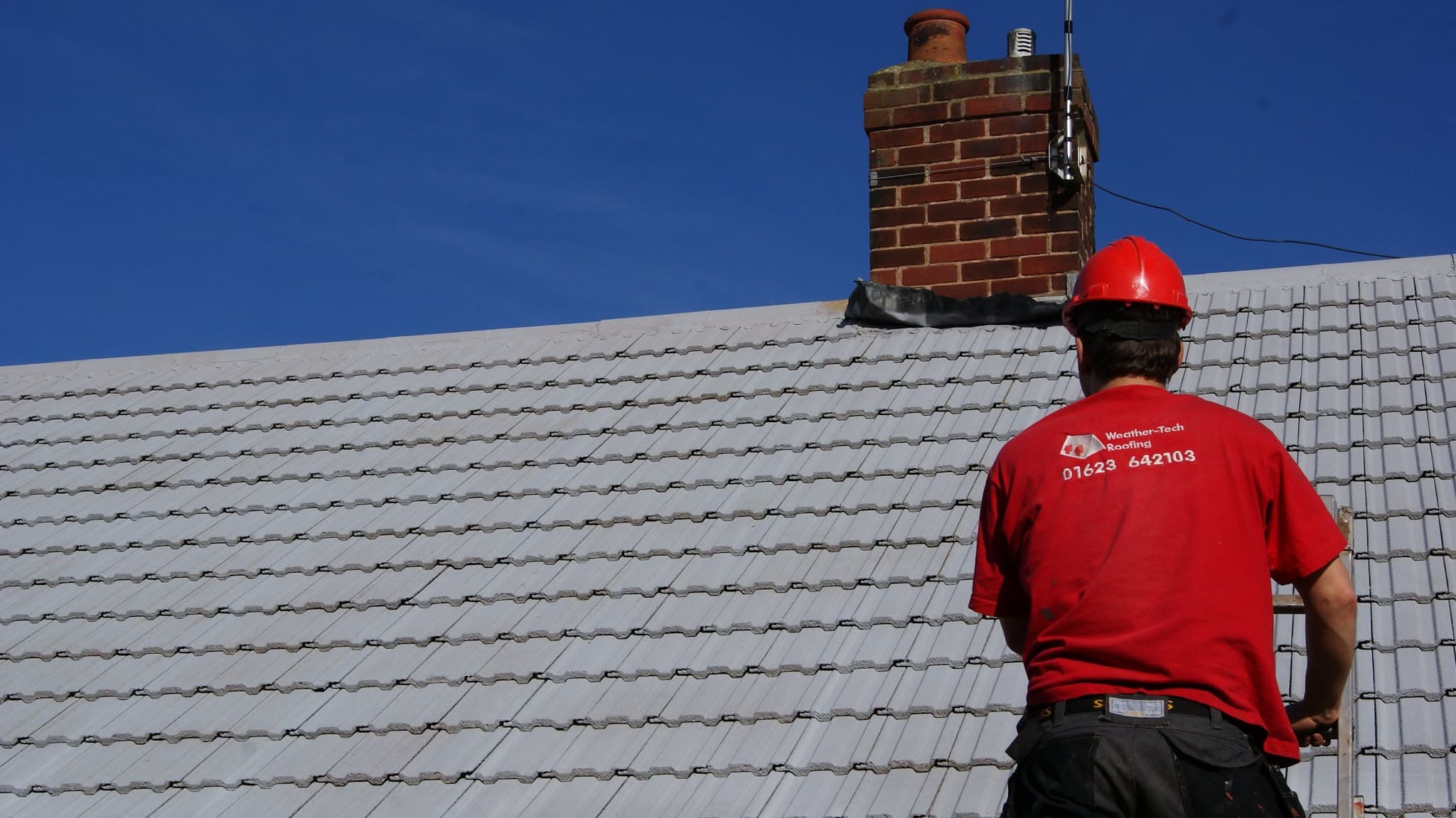 WeatherTech fitter in red hard hat inspecting grey concrete tiled pitched roof with brick chimney stack