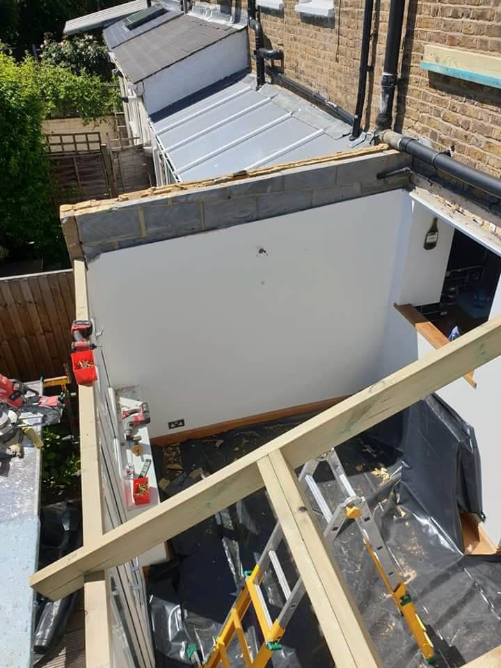 Flat roof extension under construction with timber framework and grey standing seam metal roof visible on adjacent building