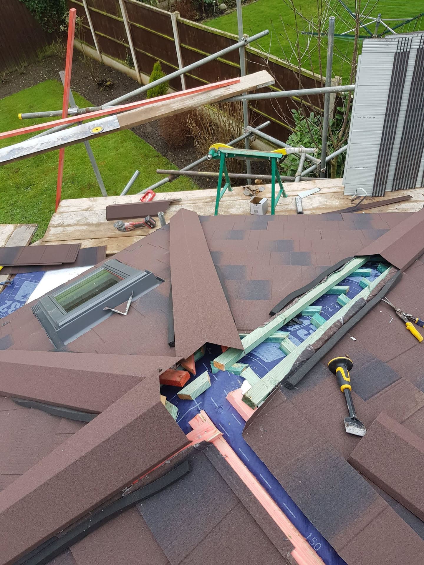 Pitched roof with brown concrete tiles being installed, showing ridge tiles, velux window and scaffolding in garden