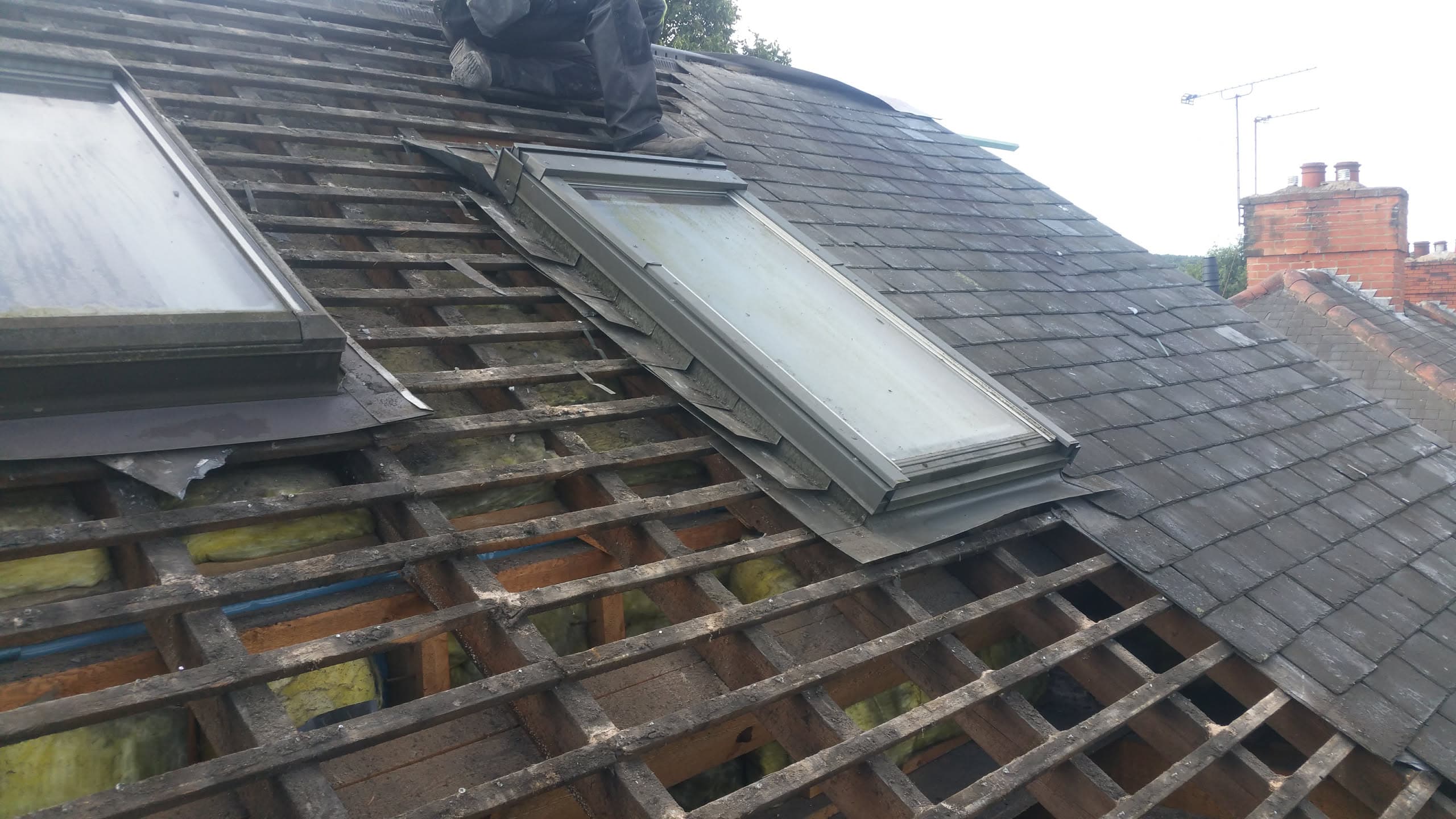 Pitched roof with exposed timber battens during slate re-roofing work, velux window visible, fitter working on ridge