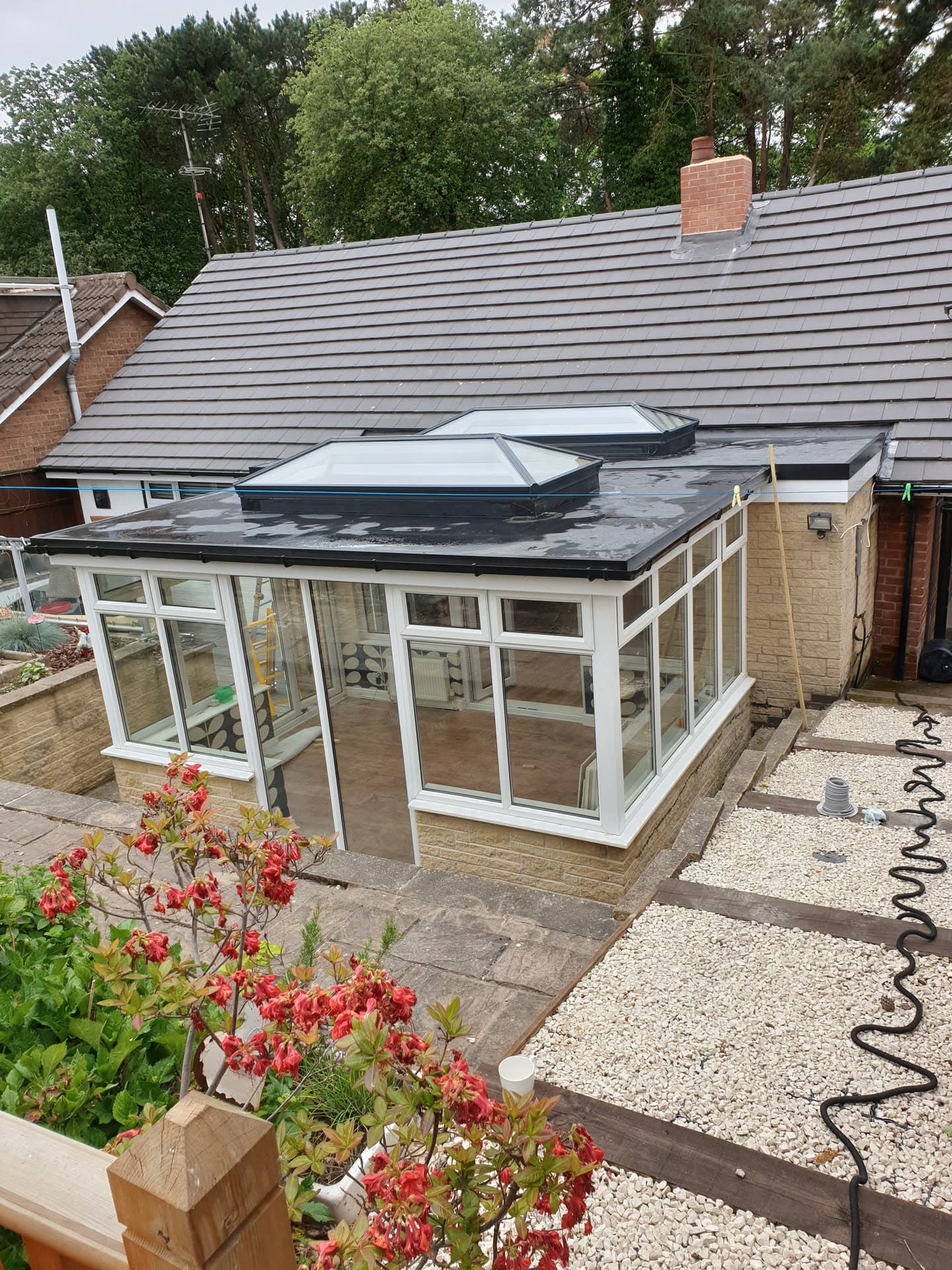 Conservatory with dark flat roof and grey lantern skylights, pitched tiled roof visible behind with brick chimney