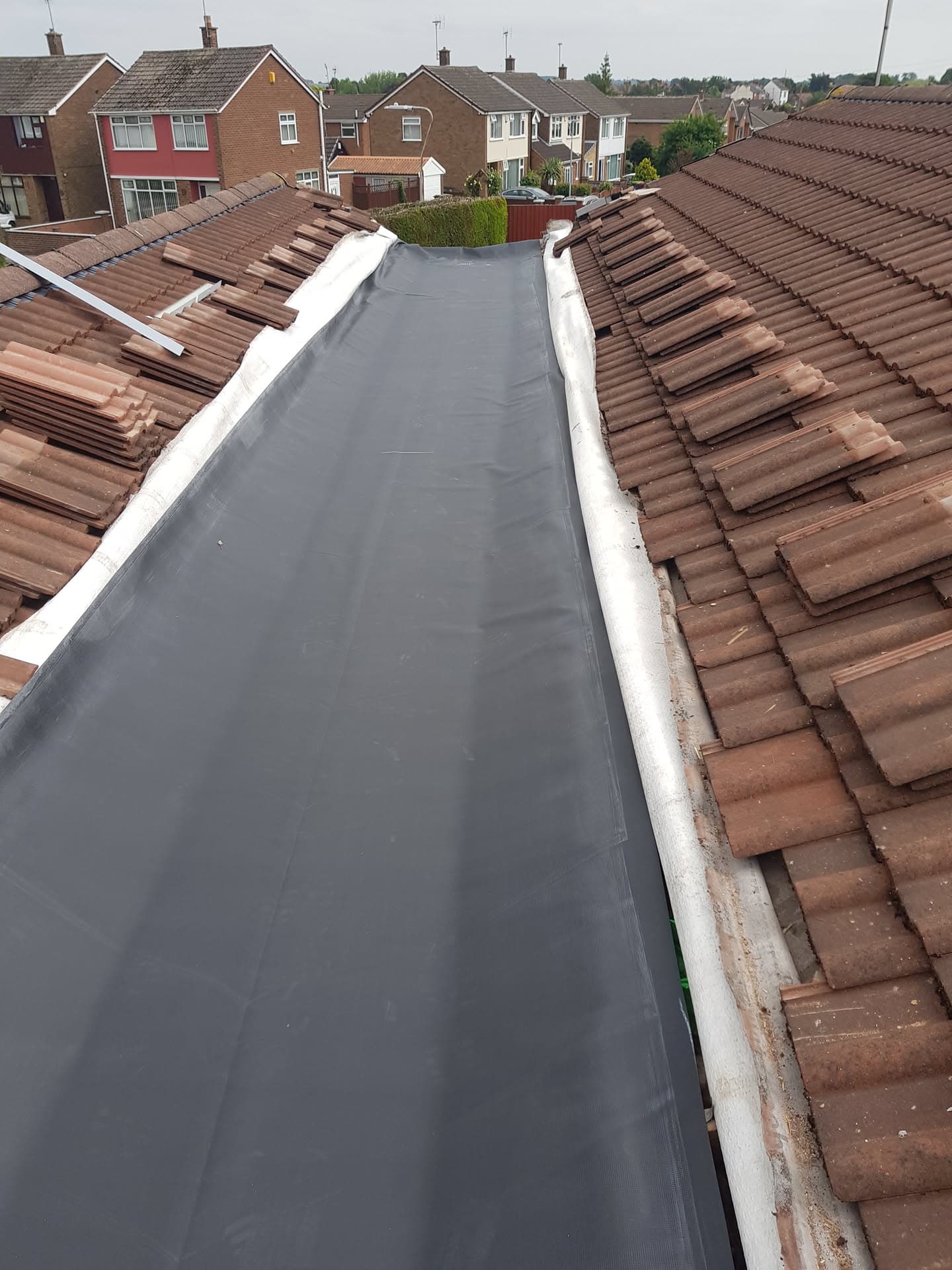 Flat roof section with grey rubber membrane between two brown tiled pitched roofs