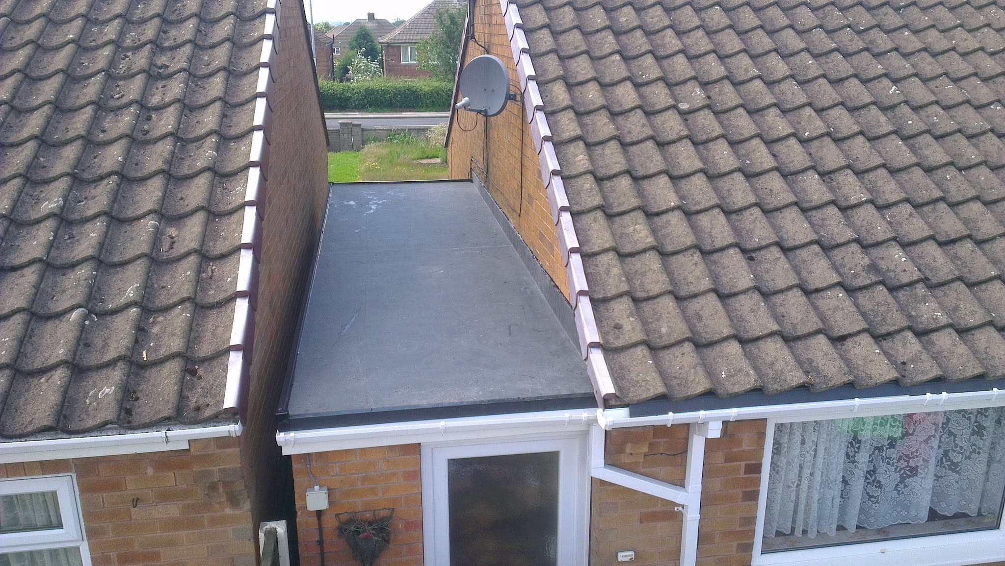 Flat roof extension with grey felt covering between two tiled pitched roofs with white fascias and guttering