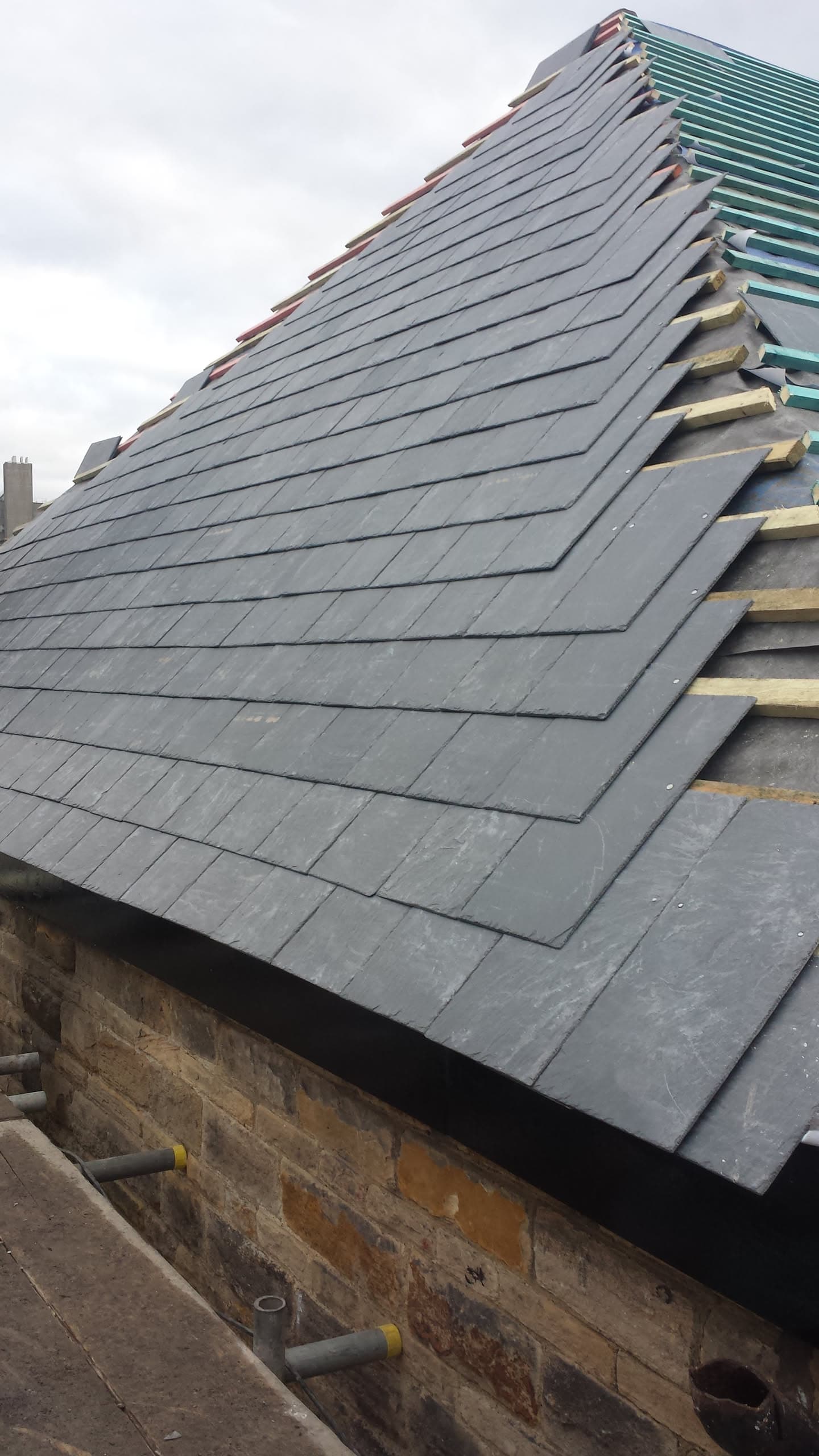 Pitched roof with grey slate tiles being installed, showing exposed battens and ridge