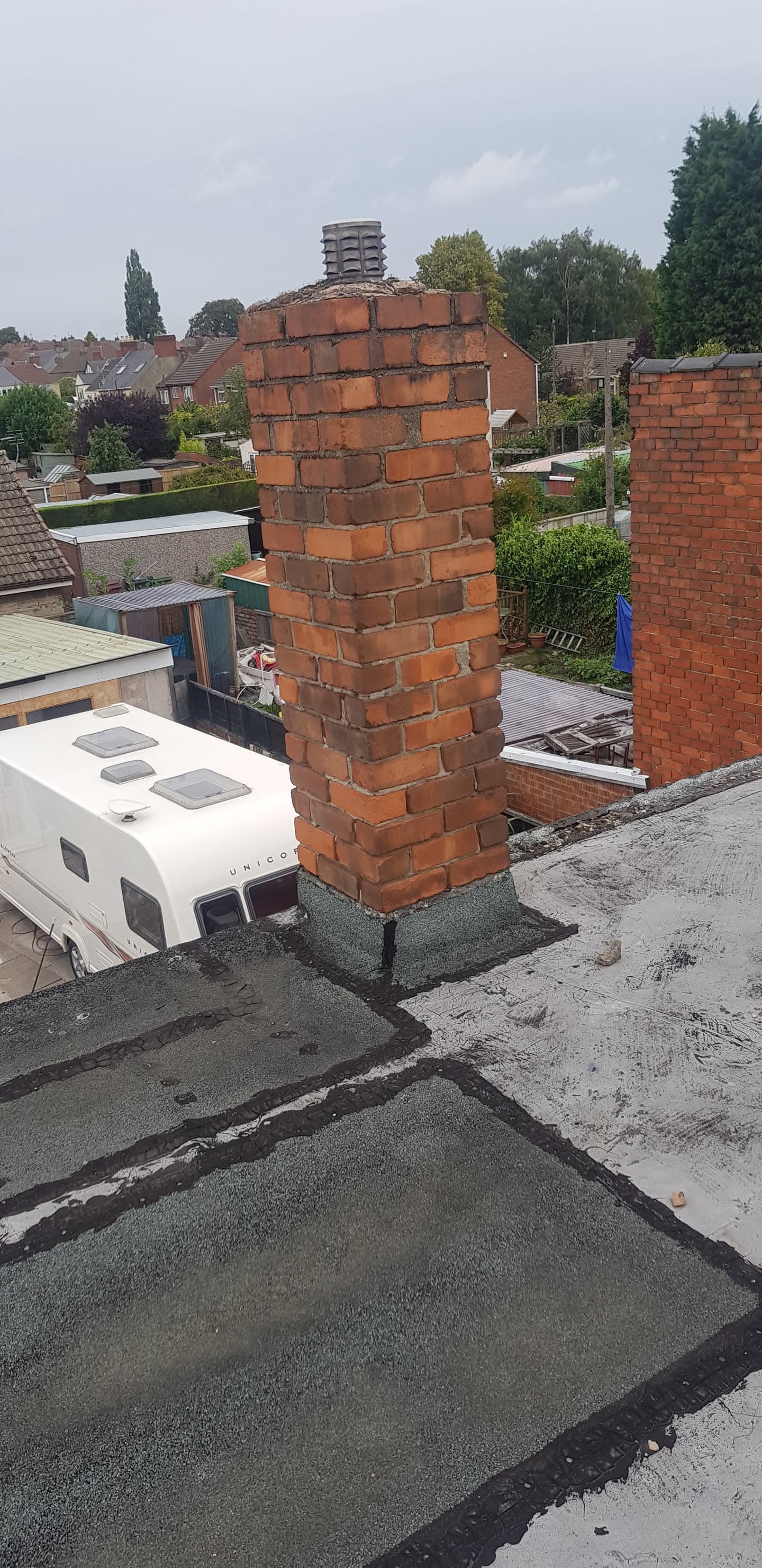 Red brick chimney stack on flat felt roof with caravan parked below