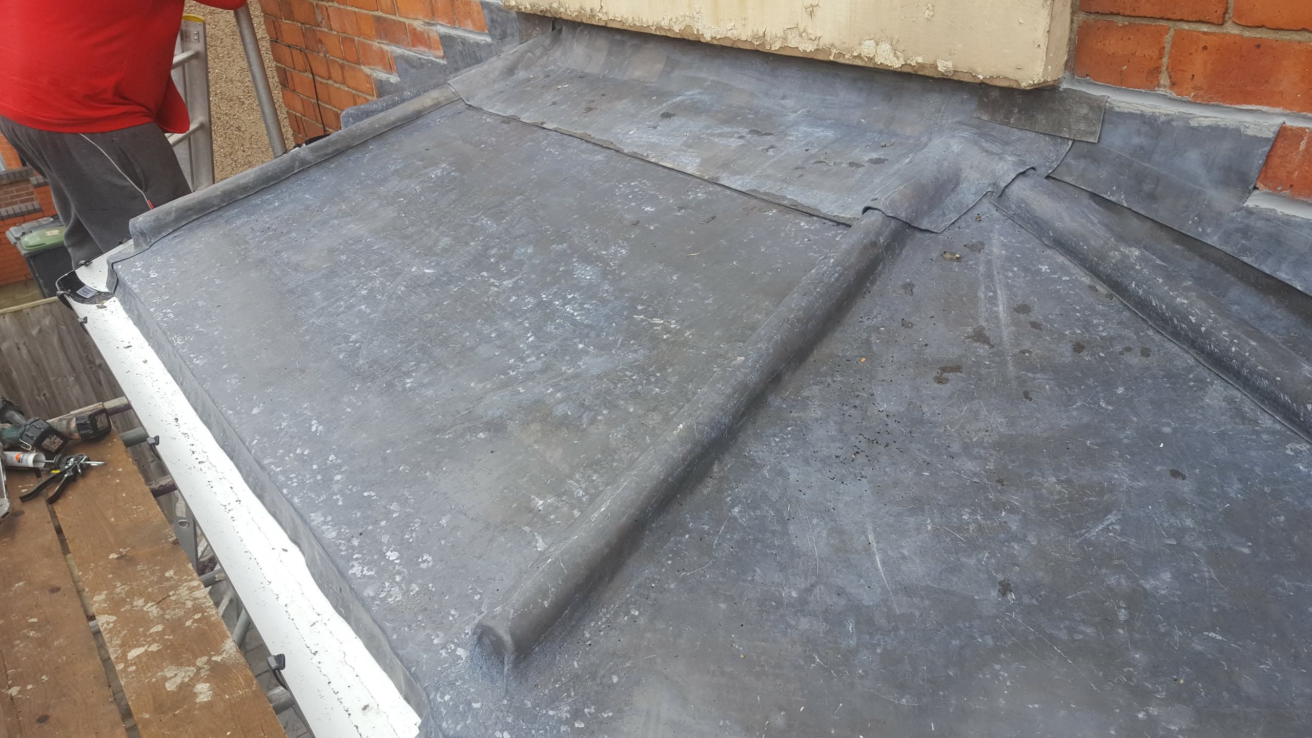 Flat roof with grey felt covering and lead flashing against brick chimney, fitter working