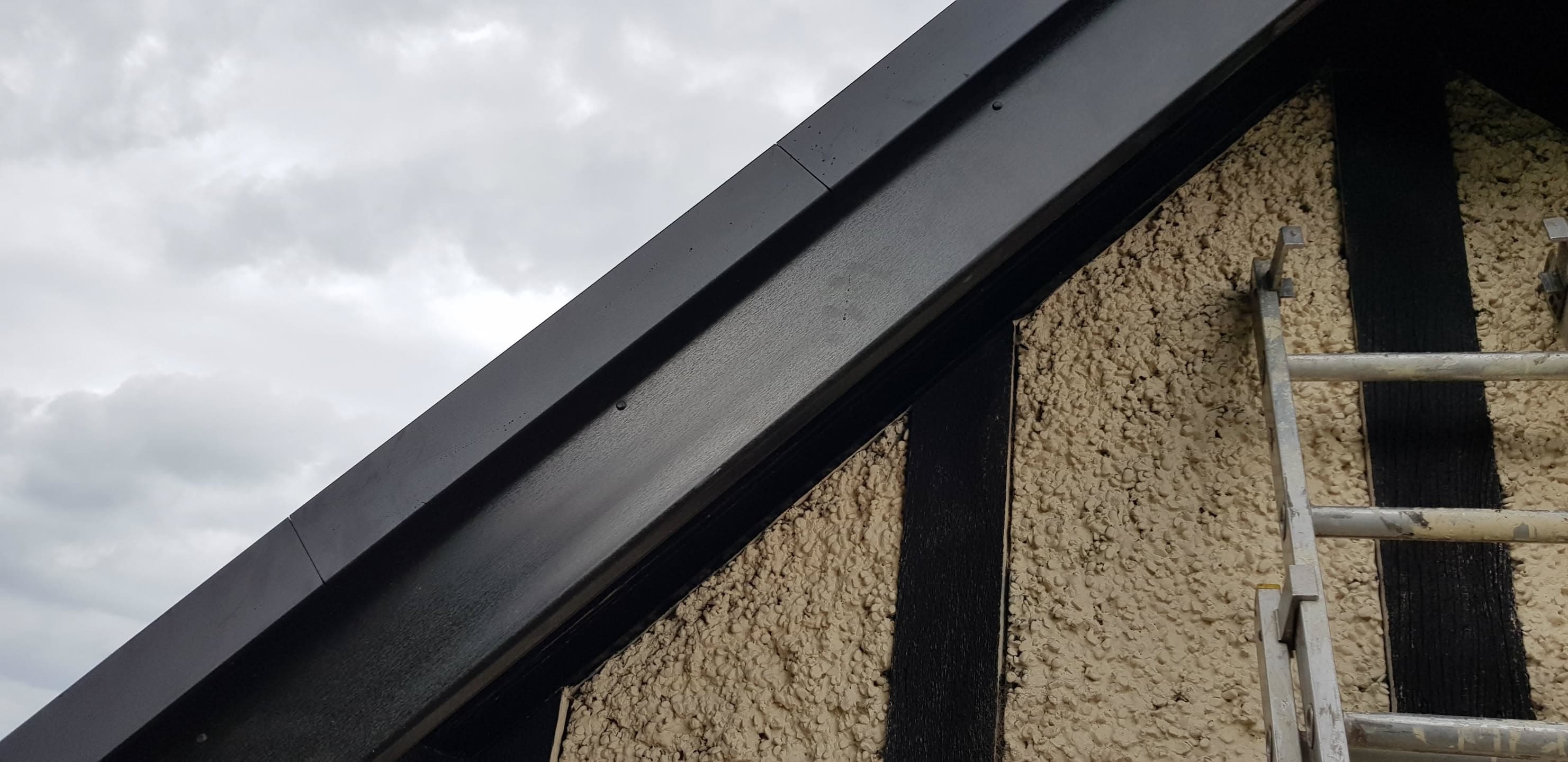 Grey metal roof edge with black fascia board and ladder against rendered wall