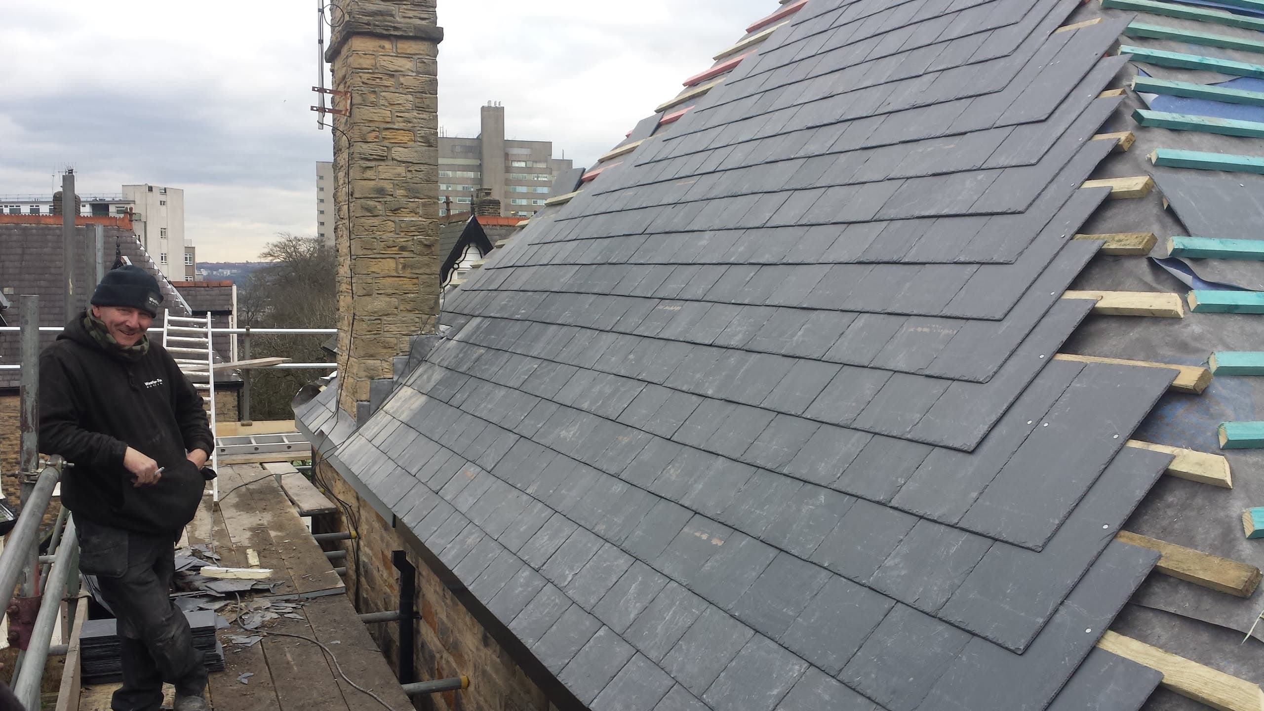 Fitter installing grey slate on pitched roof with chimney stack and scaffolding