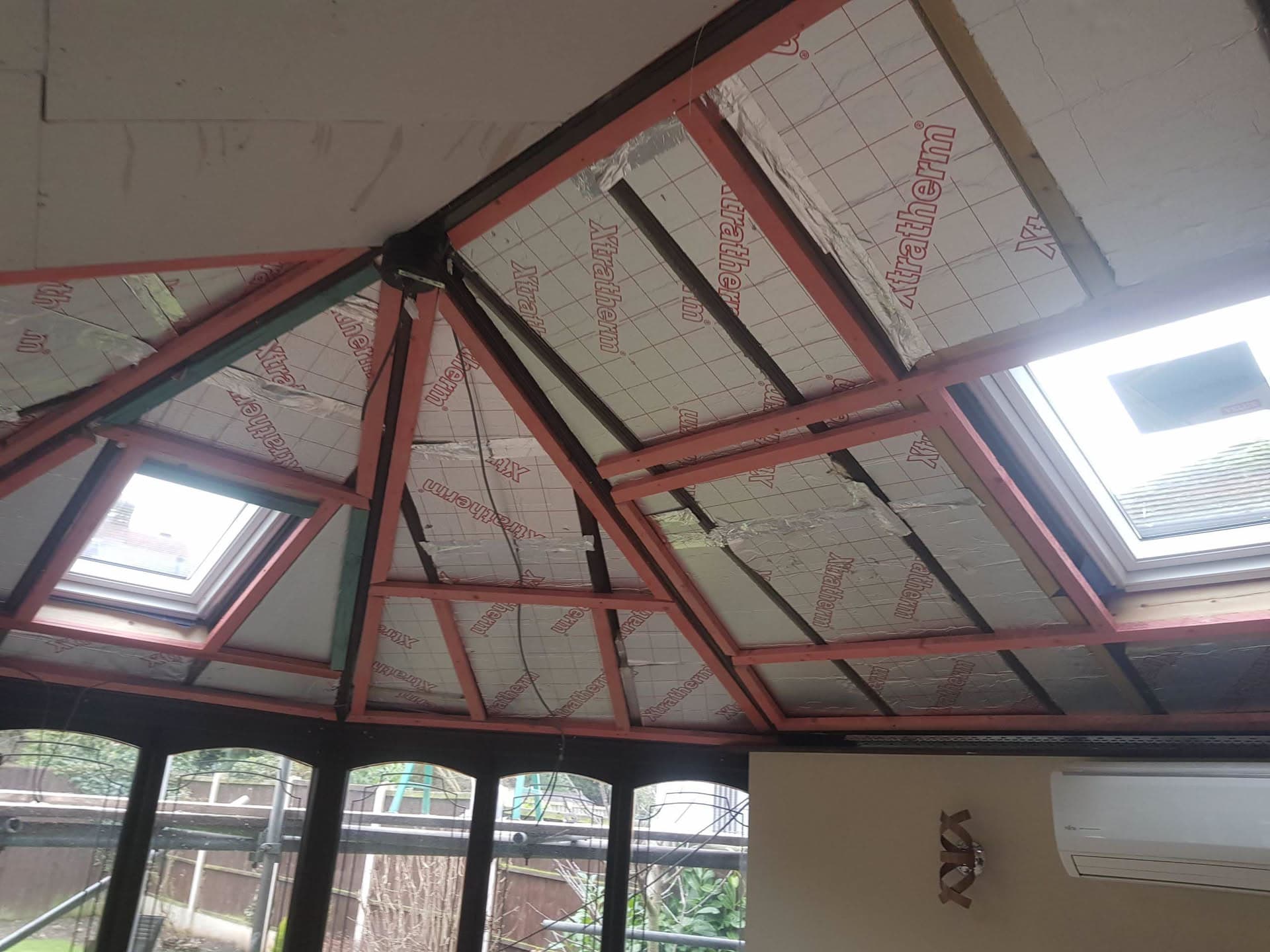 Conservatory roof interior showing insulation boards fitted between red timber rafters with skylights