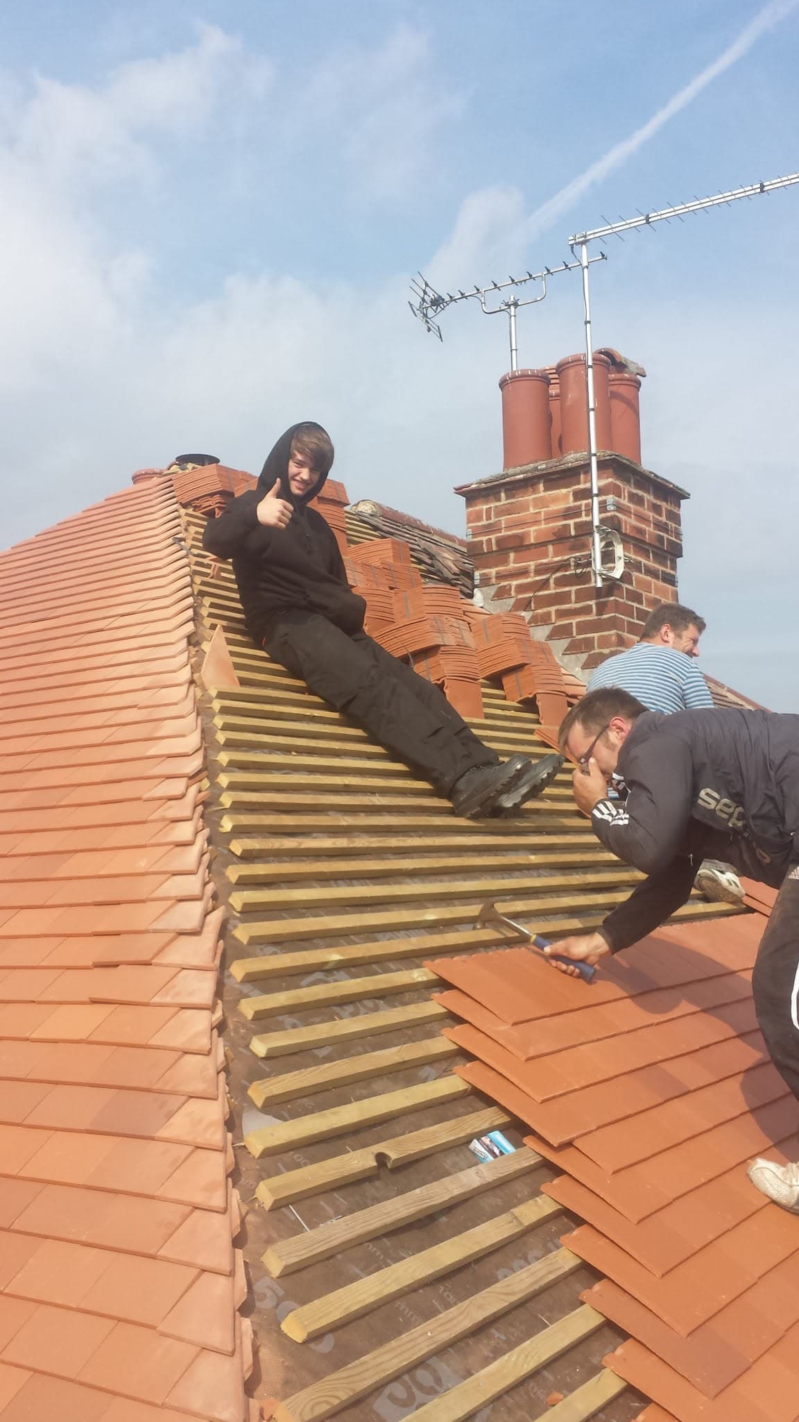 Fitters installing terracotta clay tiles on pitched roof ridge with brick chimney stack