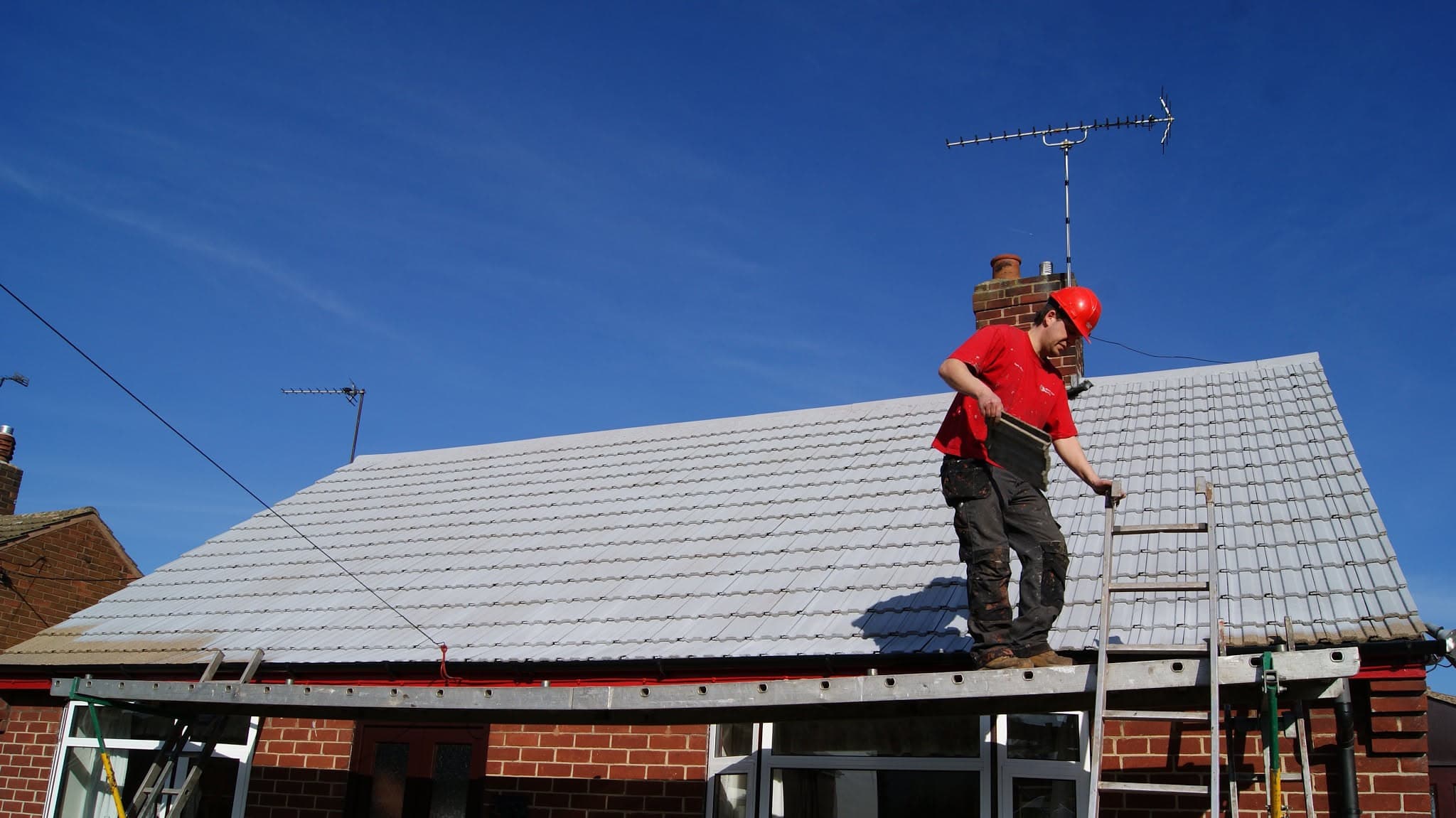 Roofer in red hard hat installing light grey concrete tiles on pitched roof with chimney and aerial
