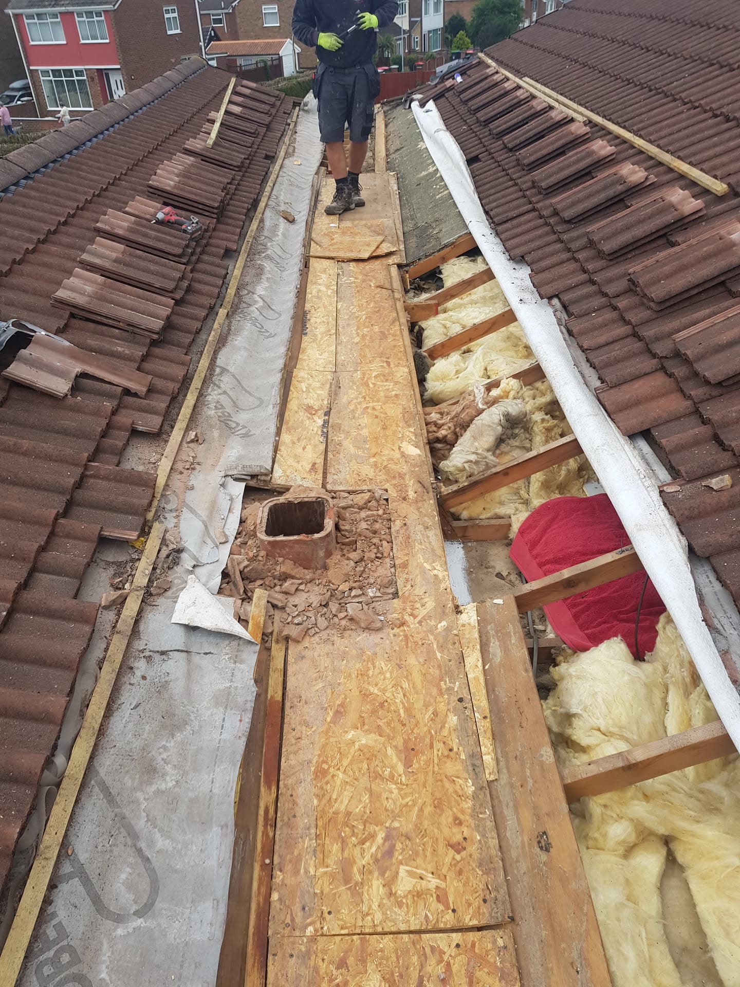 Fitter working on pitched roof with brown concrete tiles removed, exposing timber decking and insulation