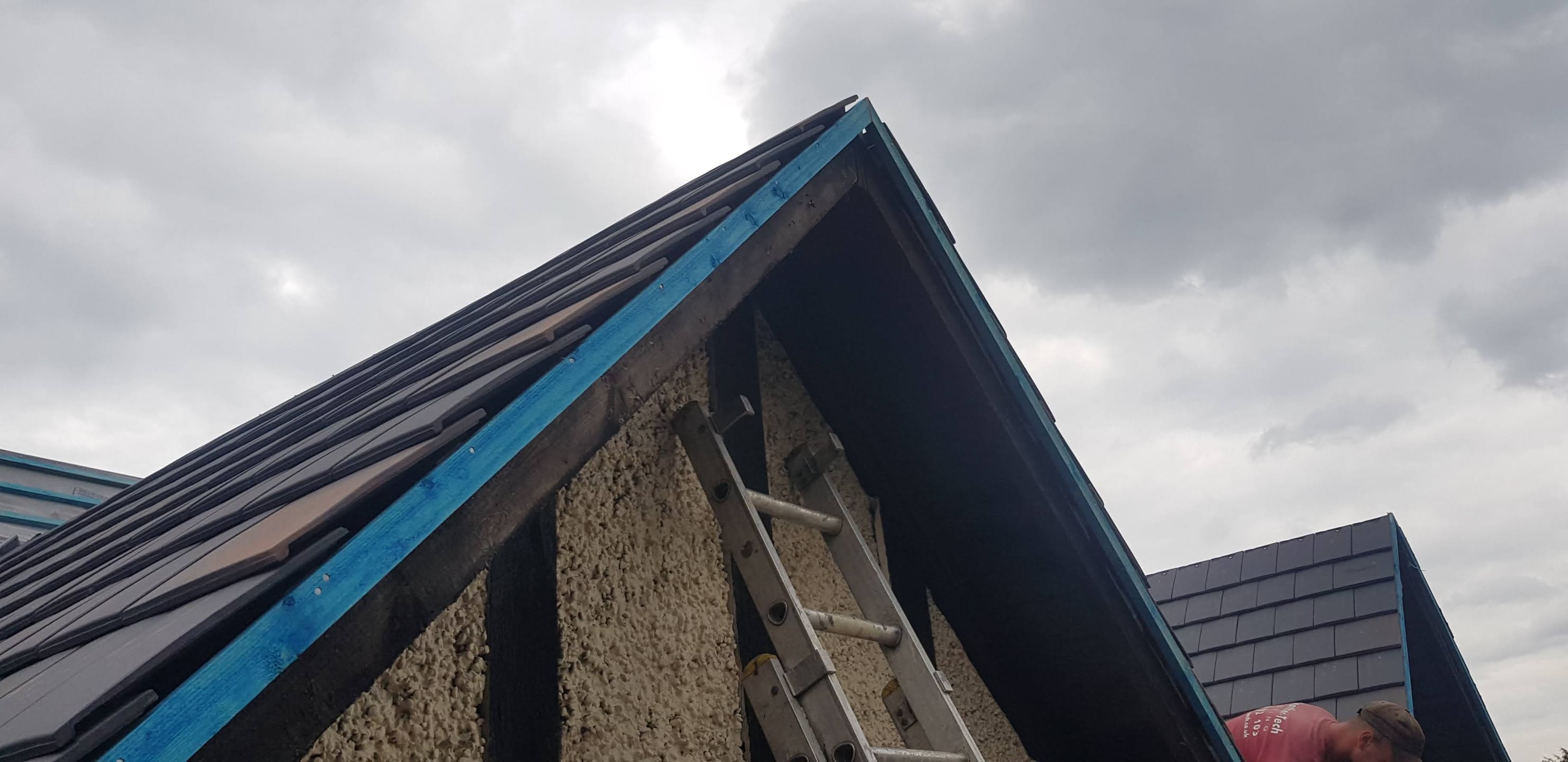 Pitched roof with dark tiles and blue fascia board with ladder against gable end