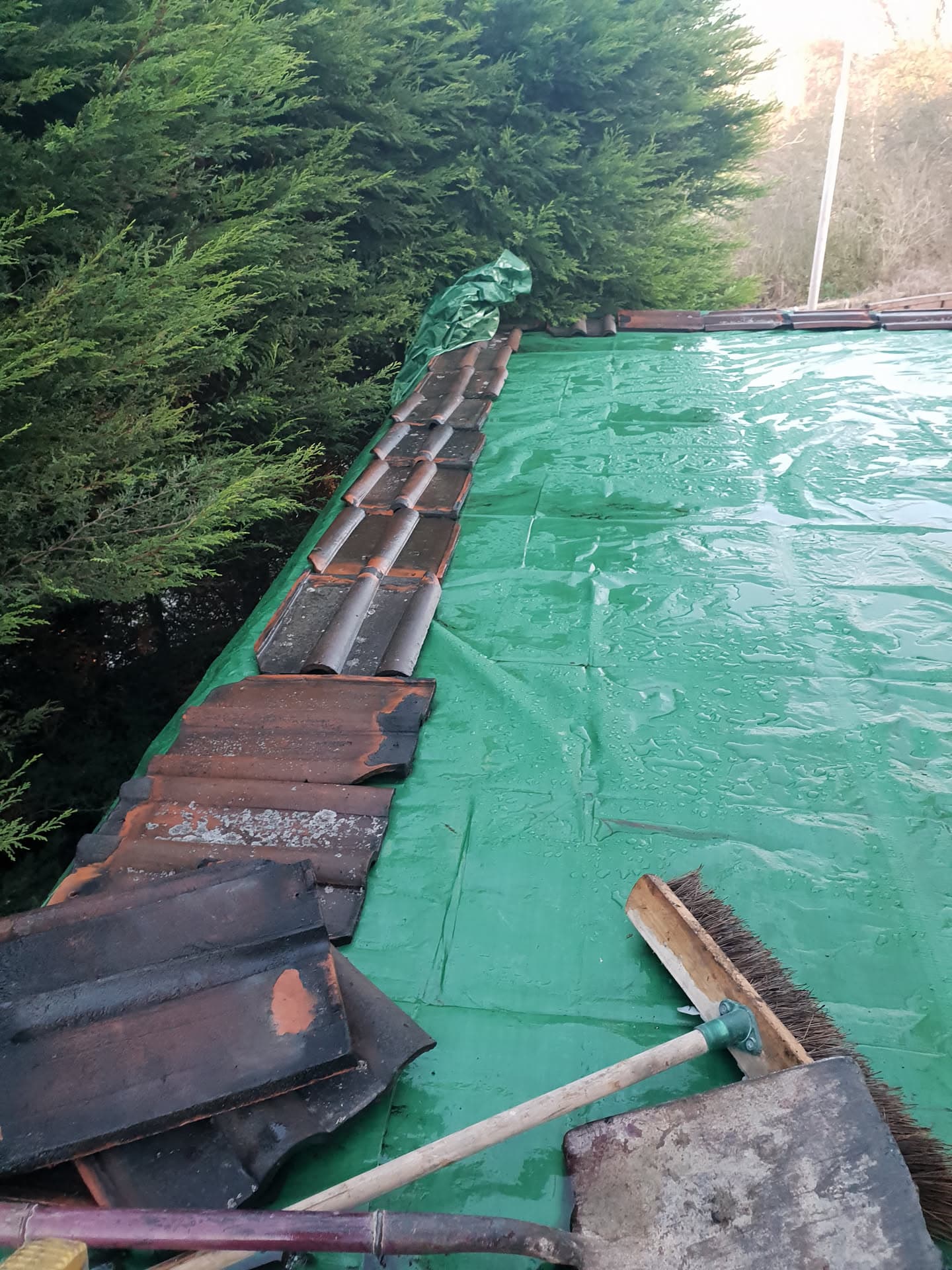 Pitched roof with dark concrete tiles being removed, green underlay visible with brush on surface