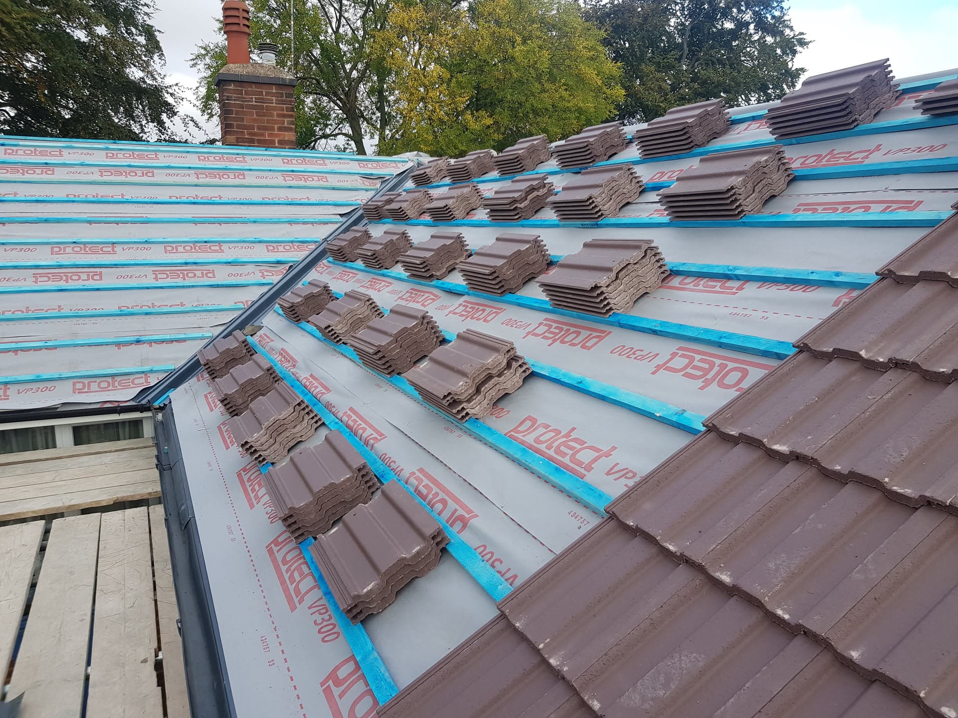 Pitched roof during re-tiling with brown concrete tiles laid on blue breathable membrane and battens, chimney visible