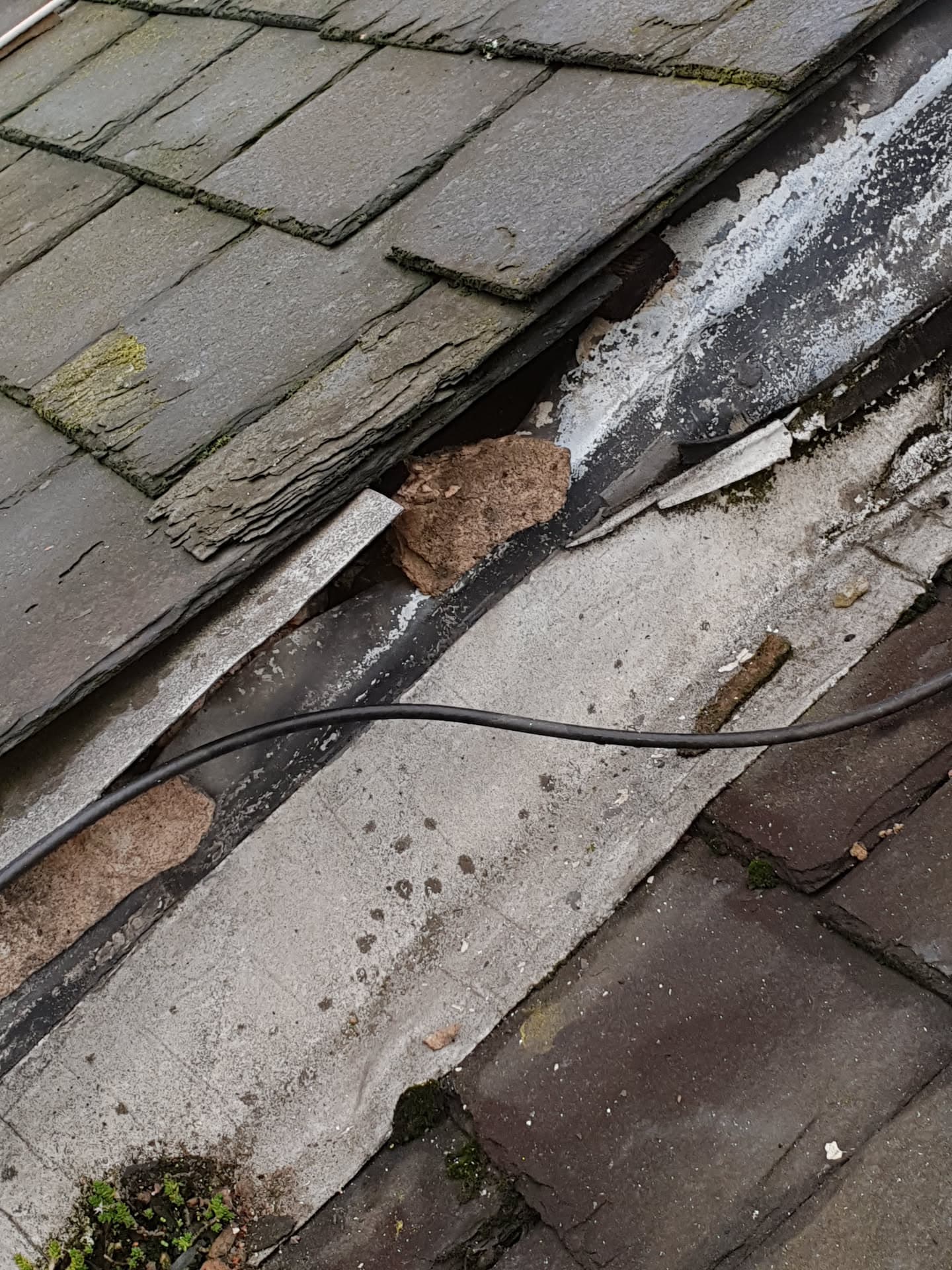 Damaged slate roof with slipped and broken slates showing deteriorated ridge and moss growth