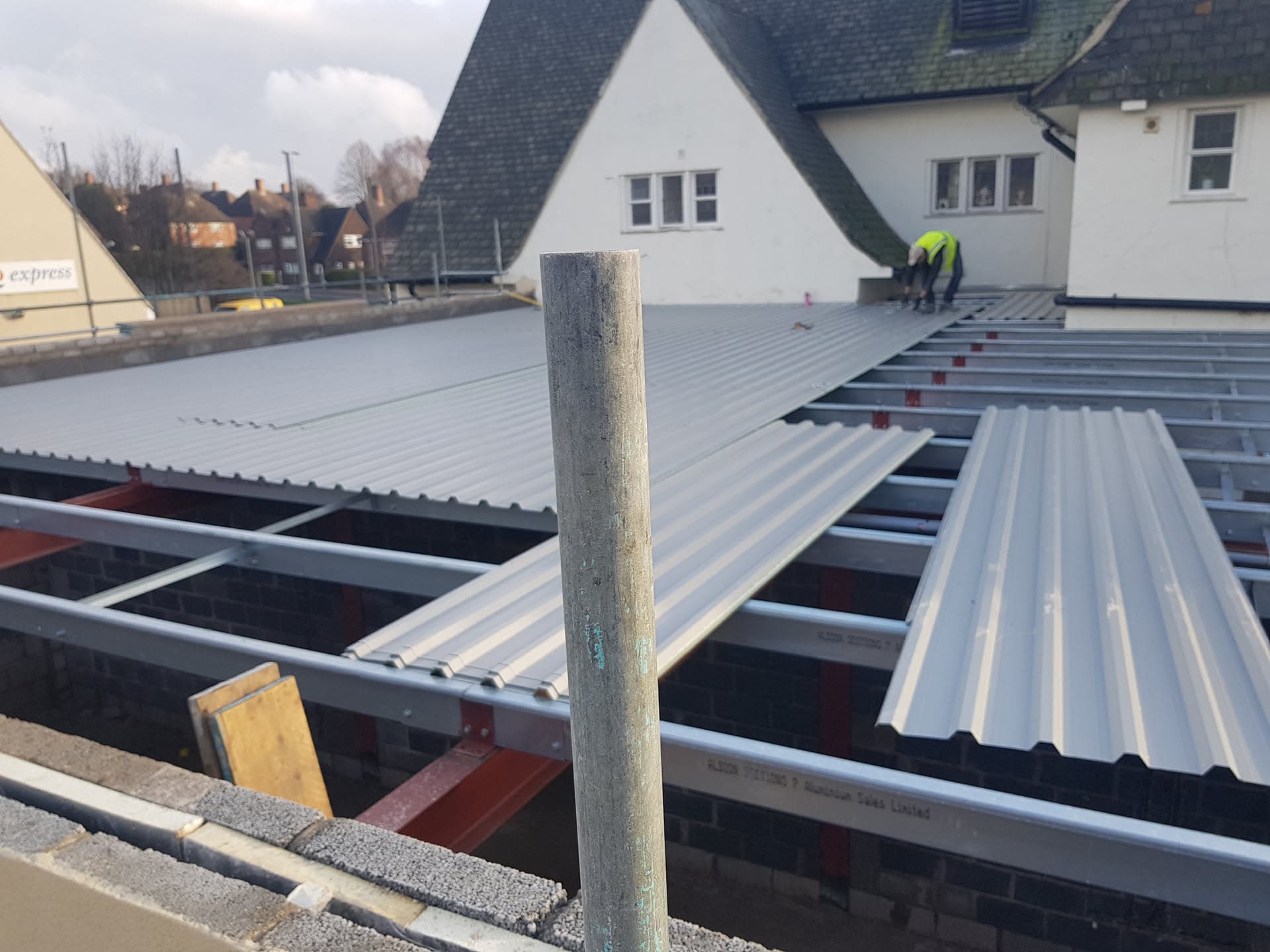Flat roof installation with grey corrugated metal sheets being fitted over steel framework, fitter working in background