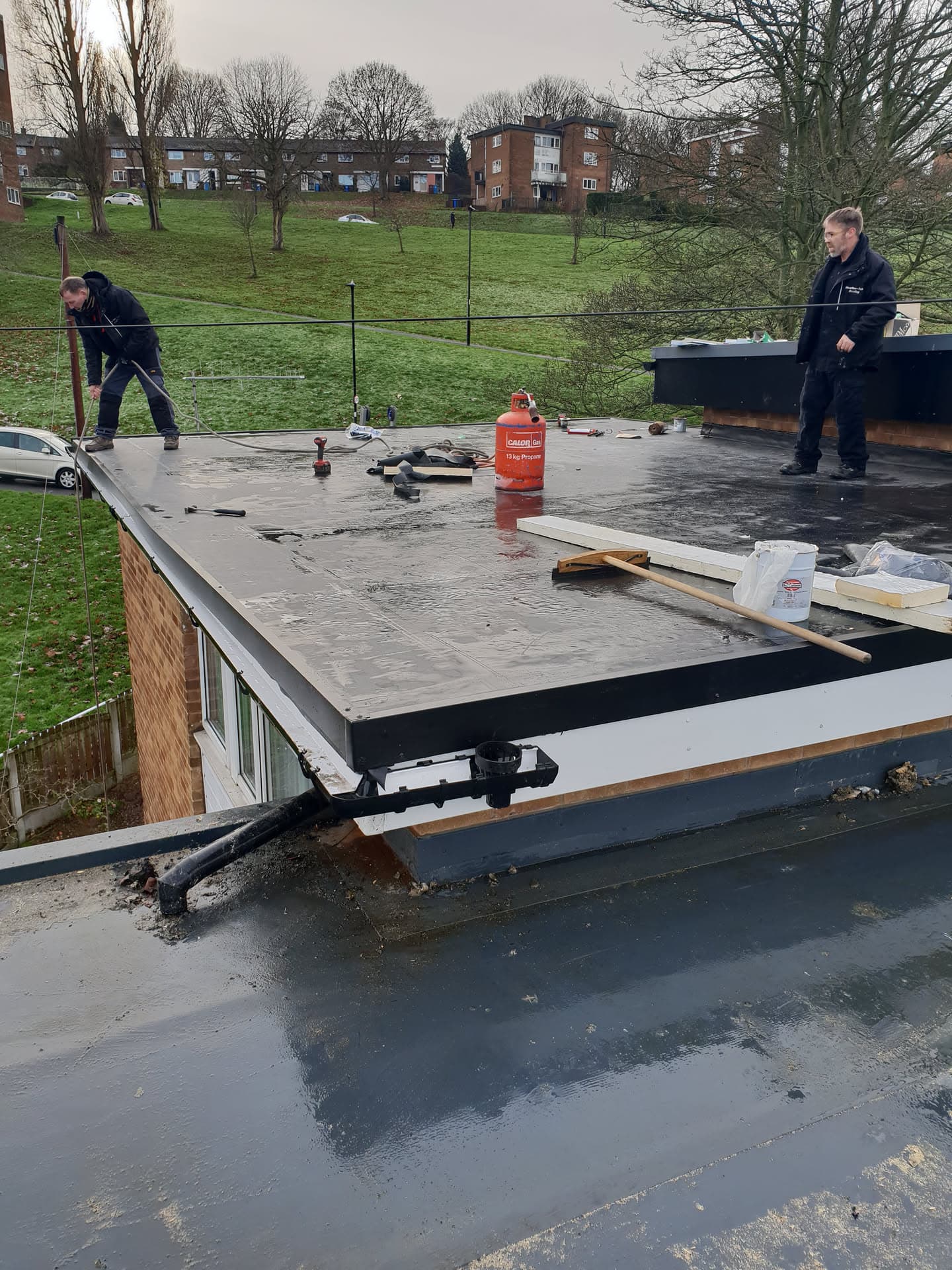 Two fitters installing grey felt flat roof on residential extension with gas torch and materials