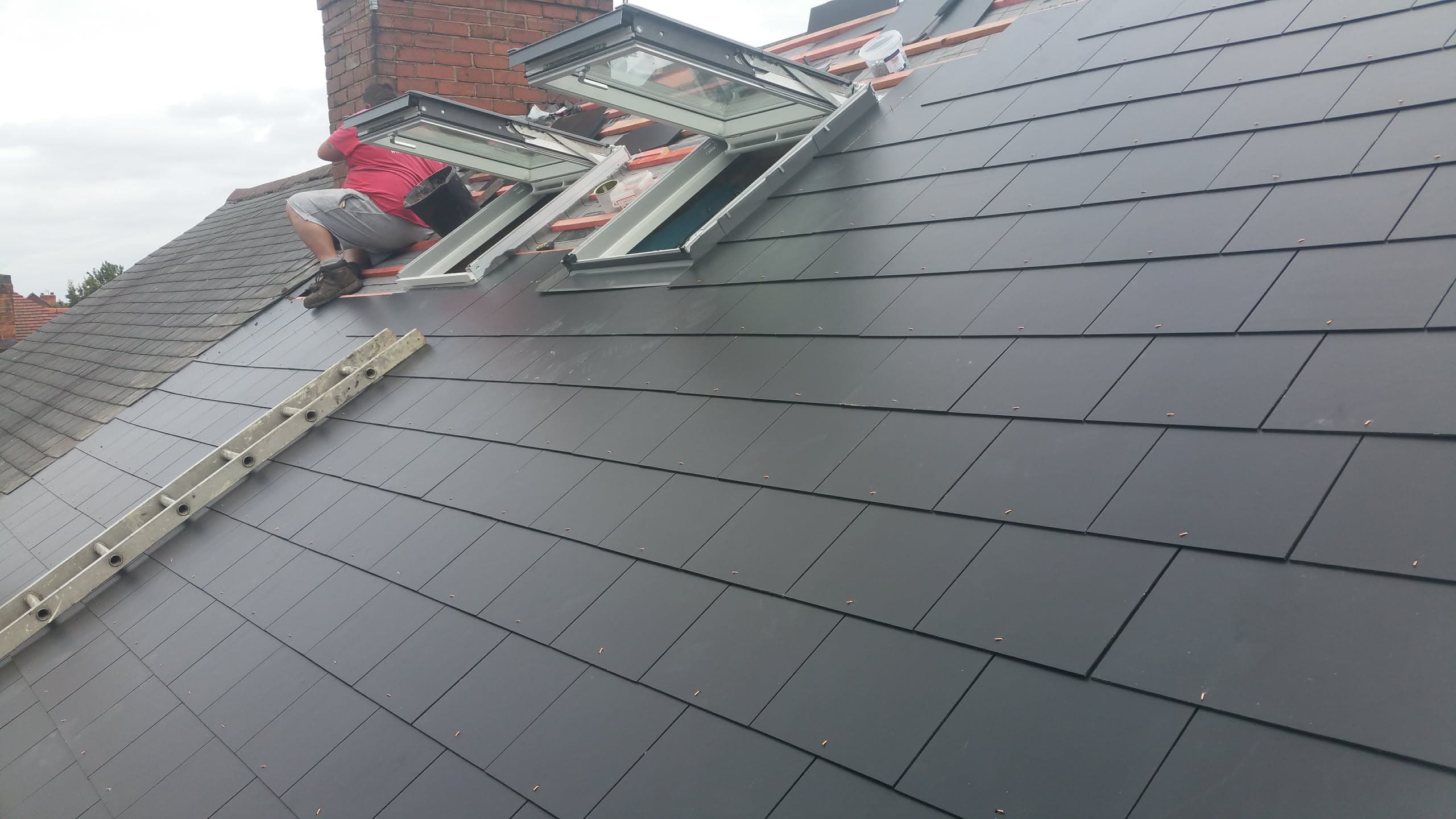 Fitter installing grey slate tiles on pitched roof with roof windows and chimney