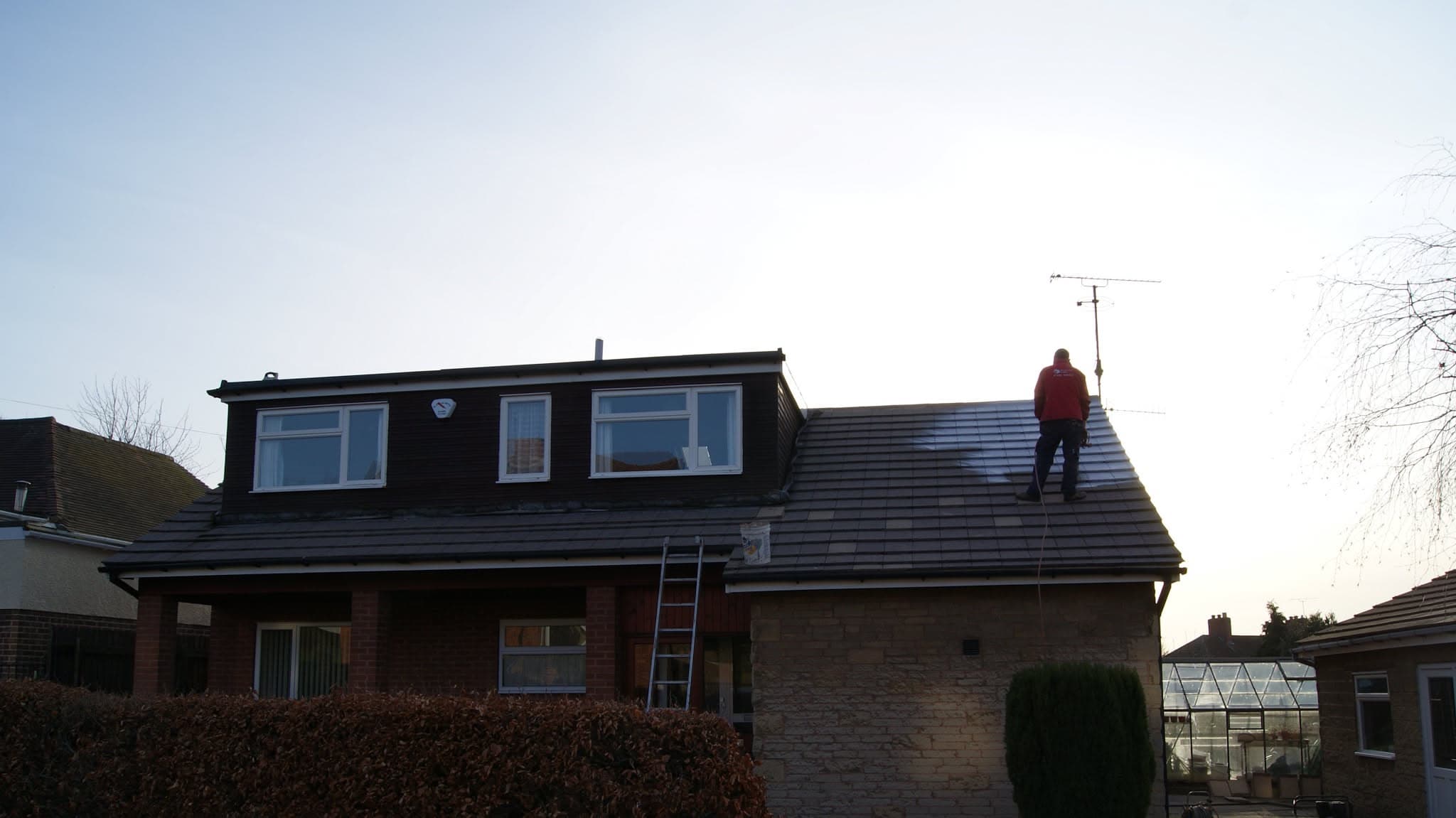 Fitter working on pitched tiled roof with ladder against brick house
