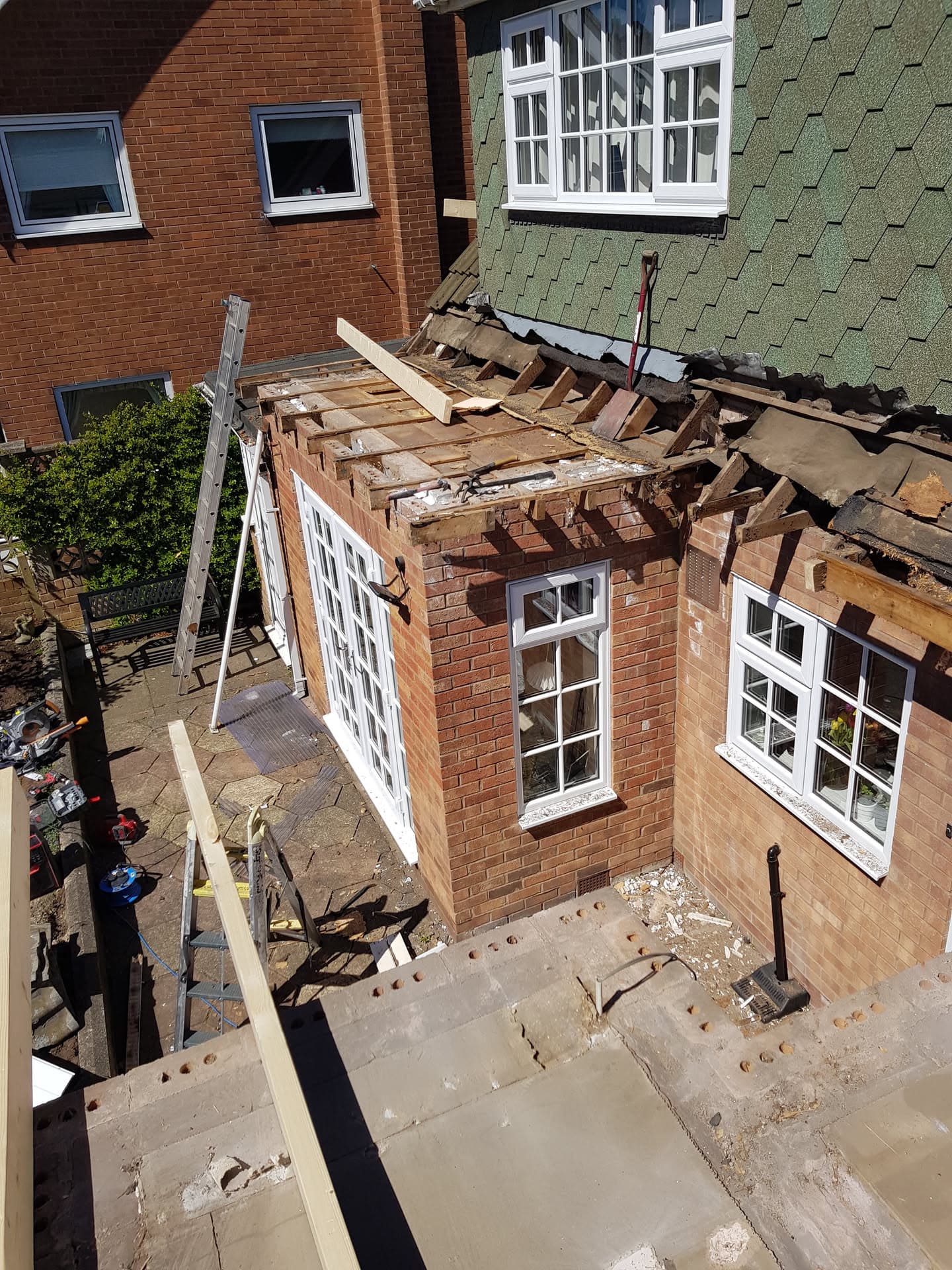 Flat roof extension being stripped and rebuilt with exposed timber joists and green hexagonal tiles on adjacent pitched roof