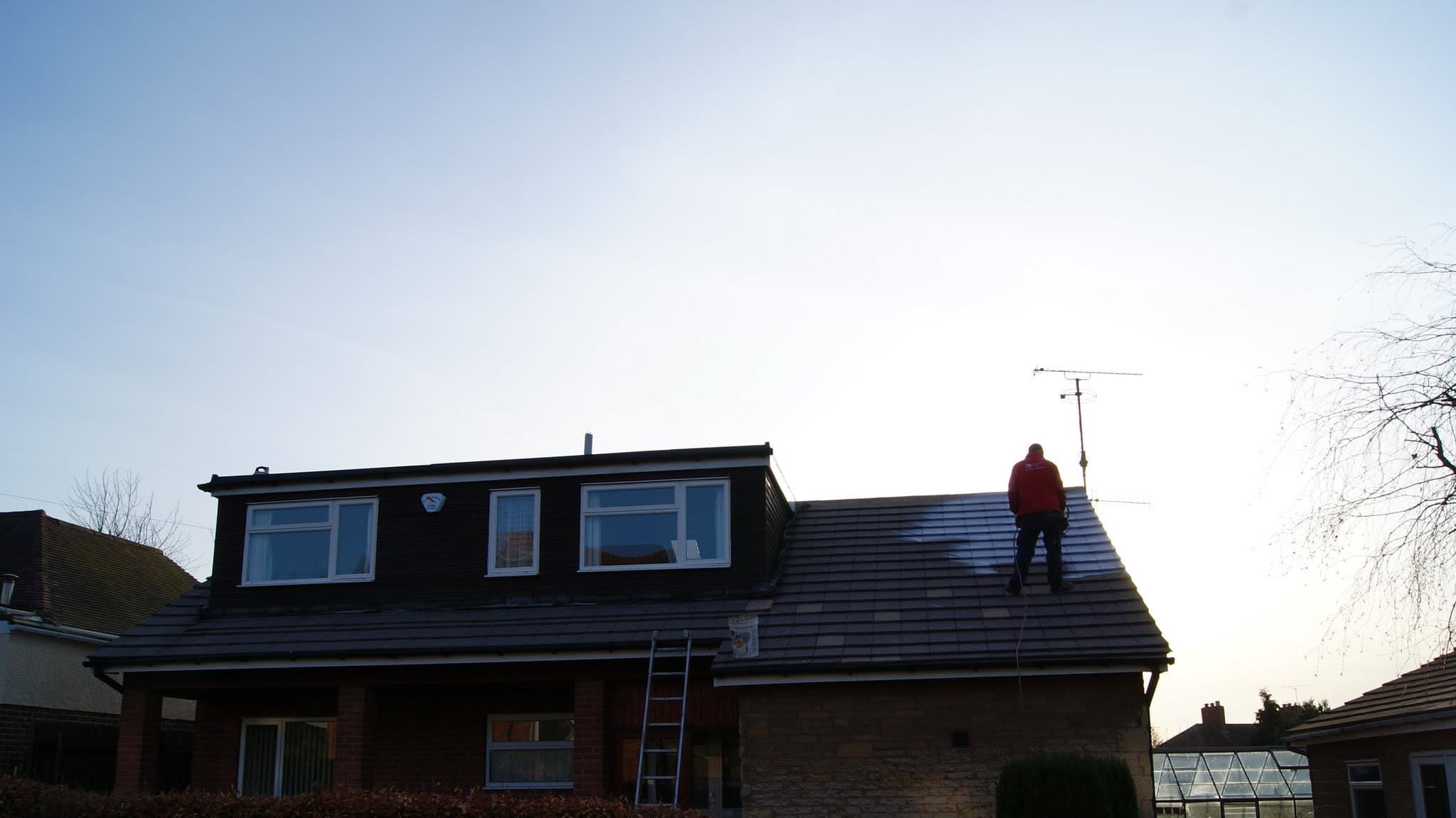 Fitter working on dark tiled pitched roof with dormer windows and ladder against property