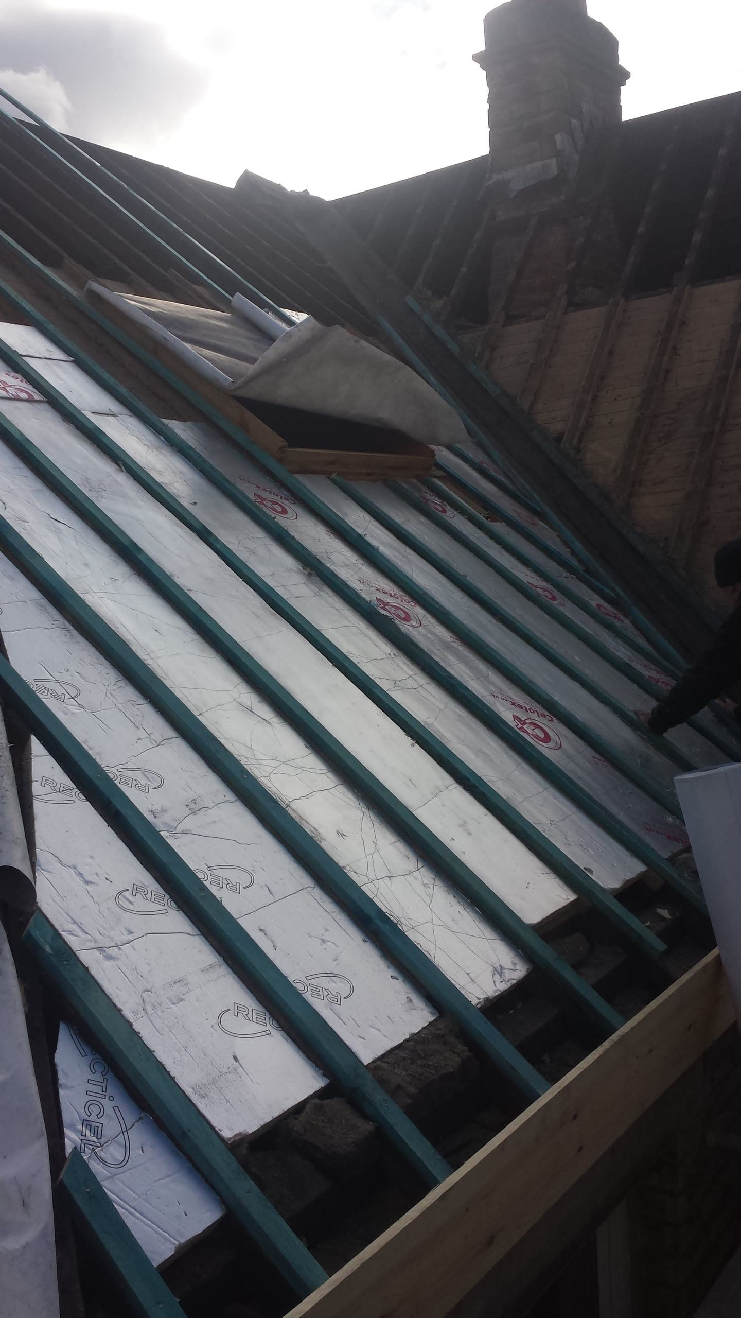 Pitched roof with dark metal standing seam panels being installed over battens and membrane, chimney visible