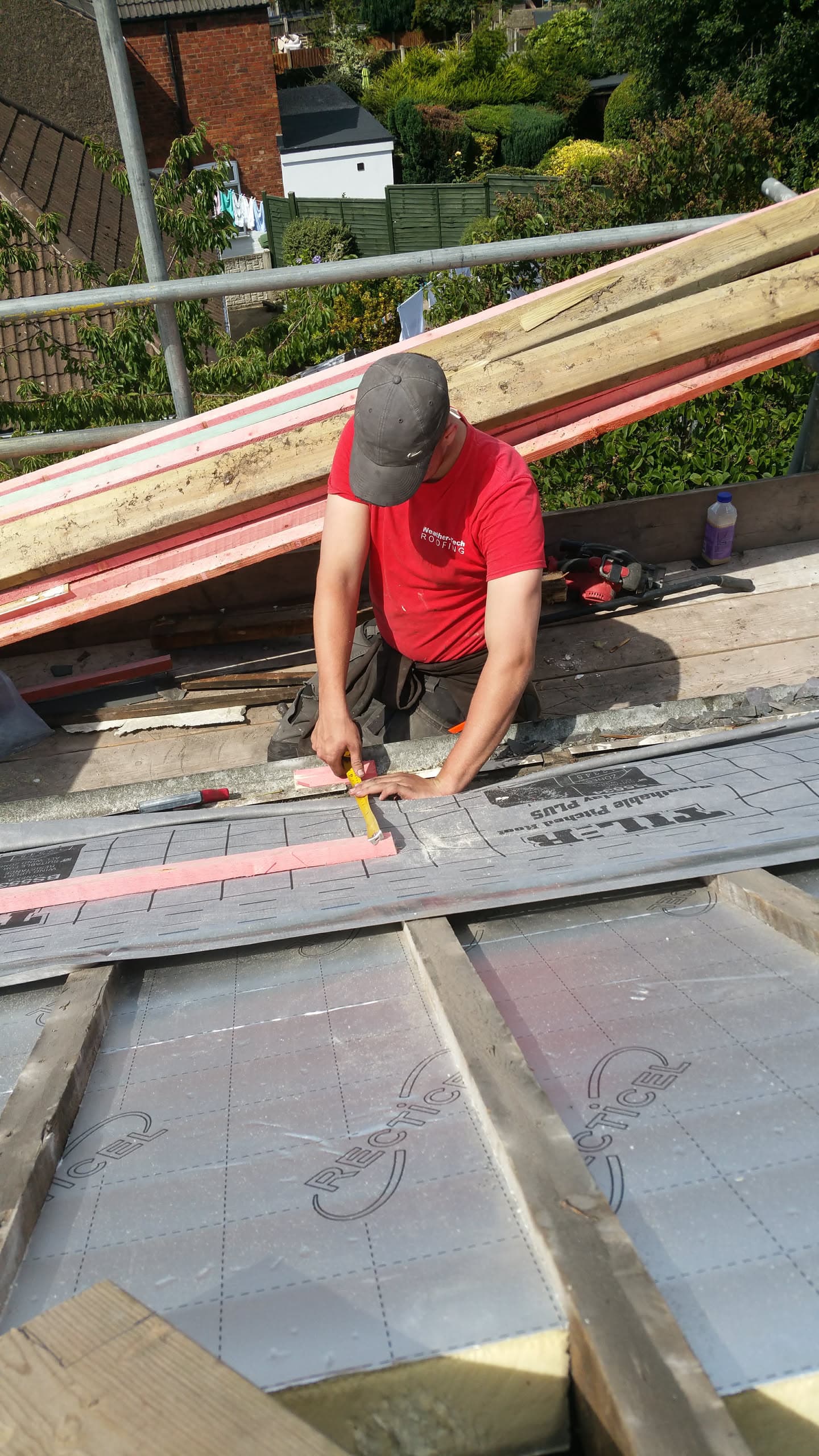 Fitter measuring grey concrete tiles on pitched roof with exposed timber battens and scaffolding
