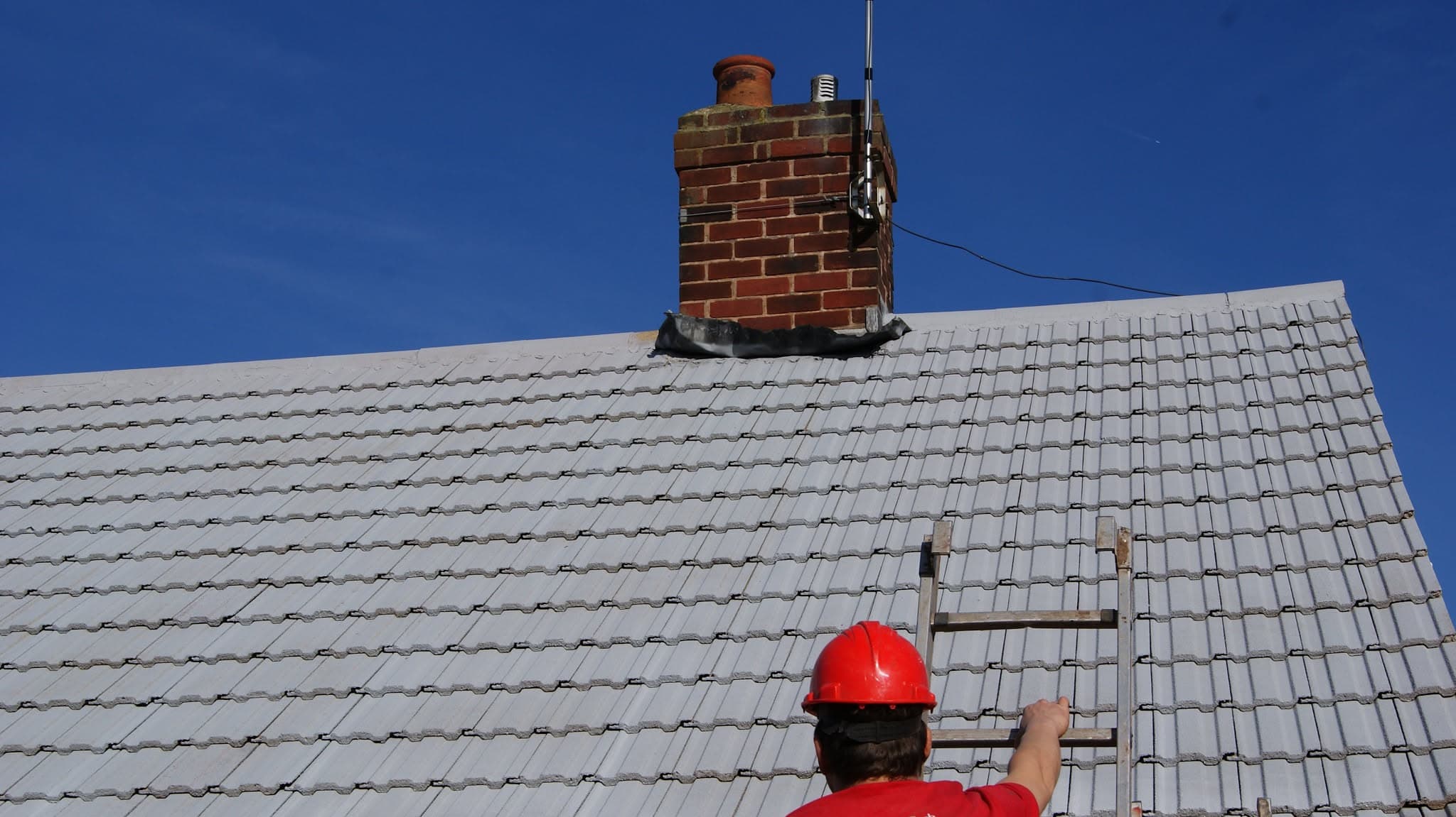 Fitter in red hard hat working on grey tiled pitched roof with brick chimney stack