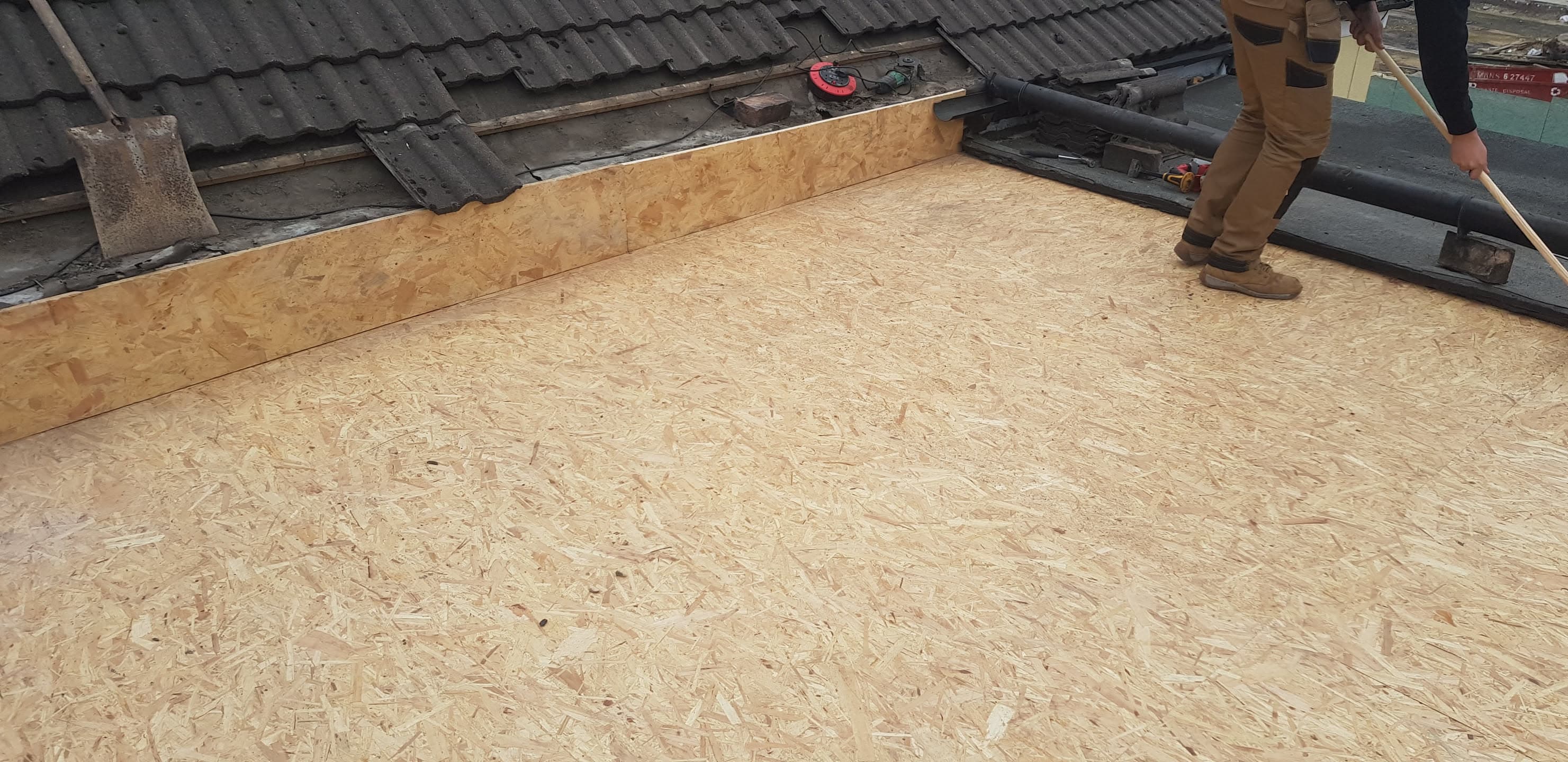 Fitter installing OSB boarding on flat roof with dark felt membrane and adjacent tiled pitched roof