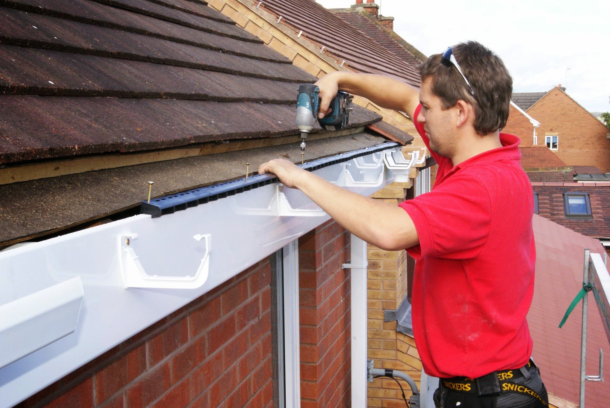 Fitter installing white fascia and guttering on pitched tiled roof using cordless drill
