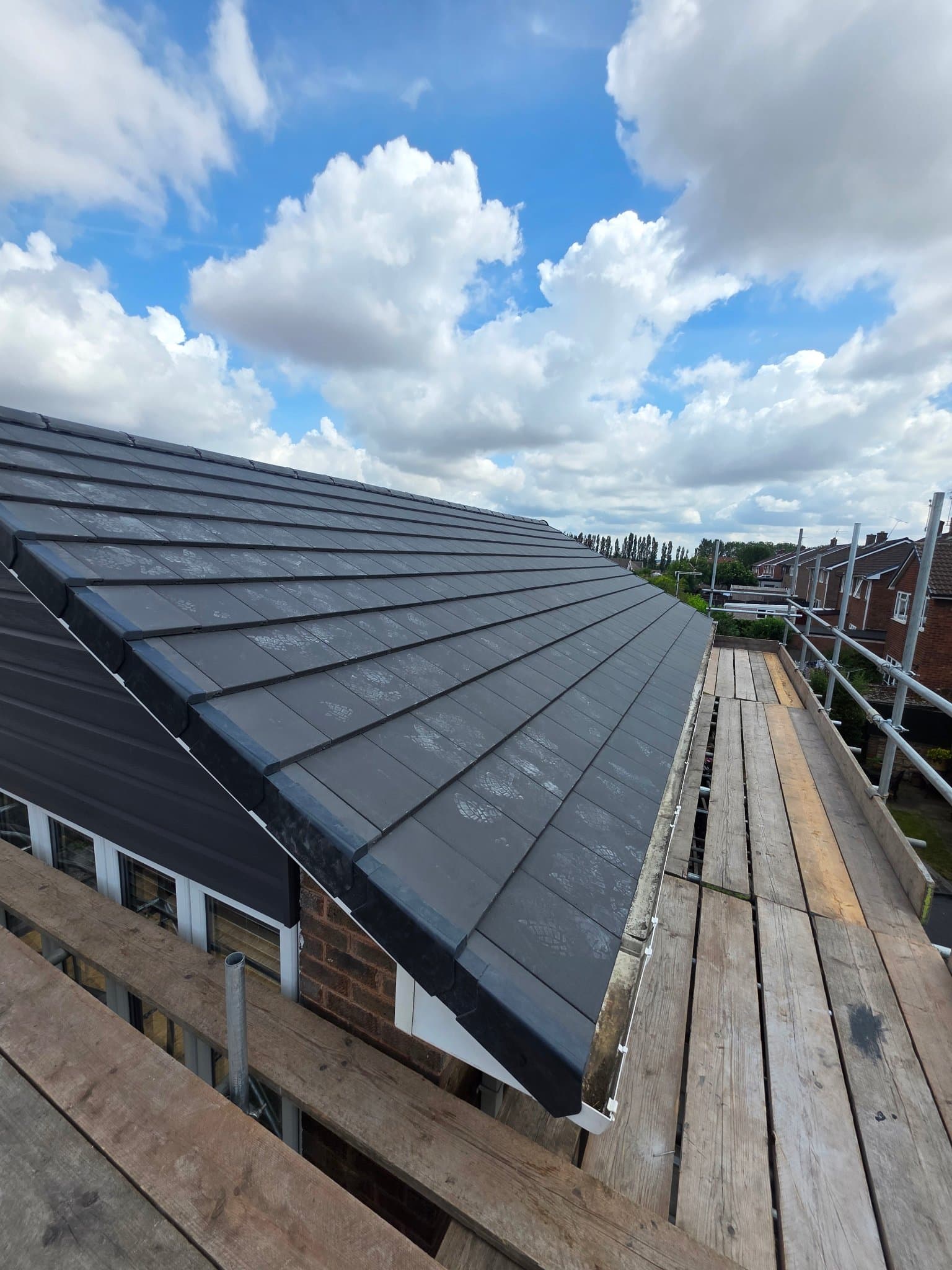 Completed dark grey tiled roof with dry verge and new guttering, scaffold still in place under a blue sky